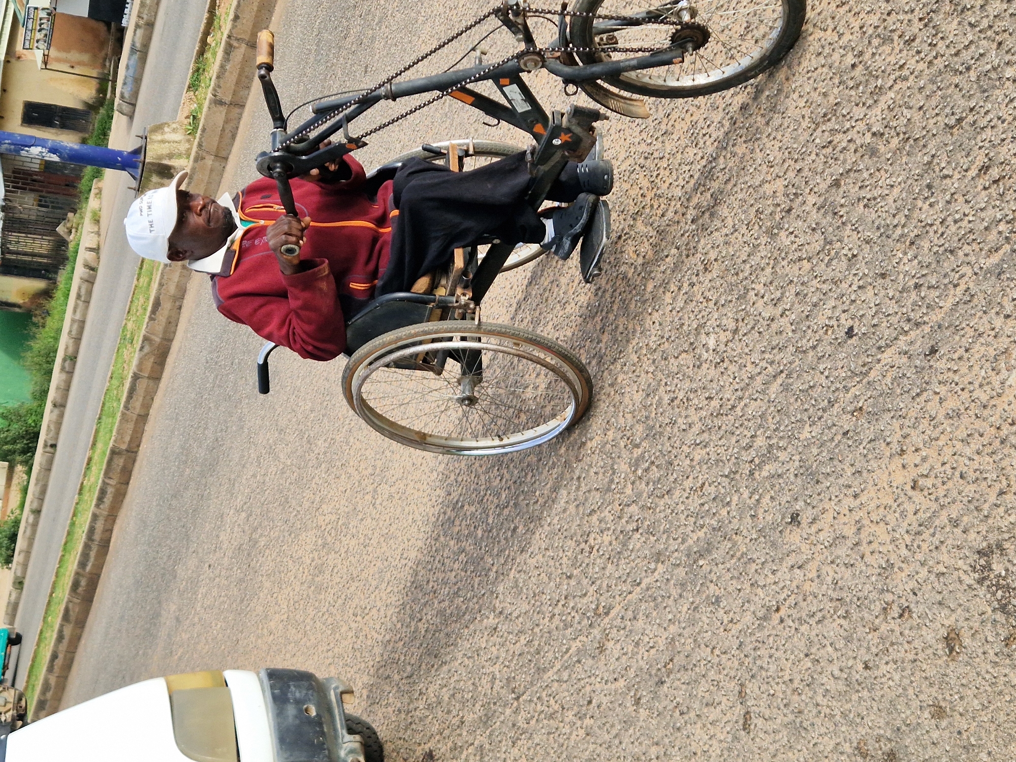 The image shows a man in a wheelchair with a modified hand-powered crank system for propulsion. He is wearing a red jacket with an orange trim, black pants, a white cap, and dark shoes. The wheelchair has large spoked rear wheels and a smaller front wheel assembly that appears to be integrated with a bicycle-like frame. The man is holding onto the hand crank mechanism. He is on a paved road with a textured asphalt surface. To the left of the man, a white vehicle is partially visible. In the background, there are buildings, a blue utility pole, and some greenery. The scene appears to be in Kabong, Nigeria, as indicated by the context. The overall lighting suggests it is daytime.