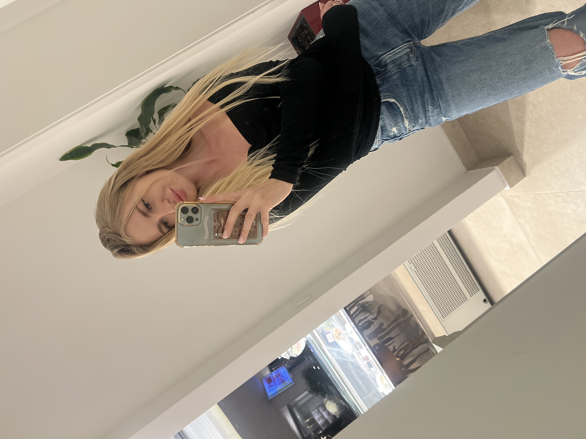 The image shows a blonde woman in a black long-sleeve shirt and ripped blue jeans taking a selfie in what appears to be a home interior. She is holding a phone with a light blue case. Behind her is a white wall with a plant in the corner, a white countertop, and a glass display case with food items visible inside. A television is visible in the background. The scene is well-lit with a tiled floor. There is no information in the image to confirm it is Boreti, Montenegro.
