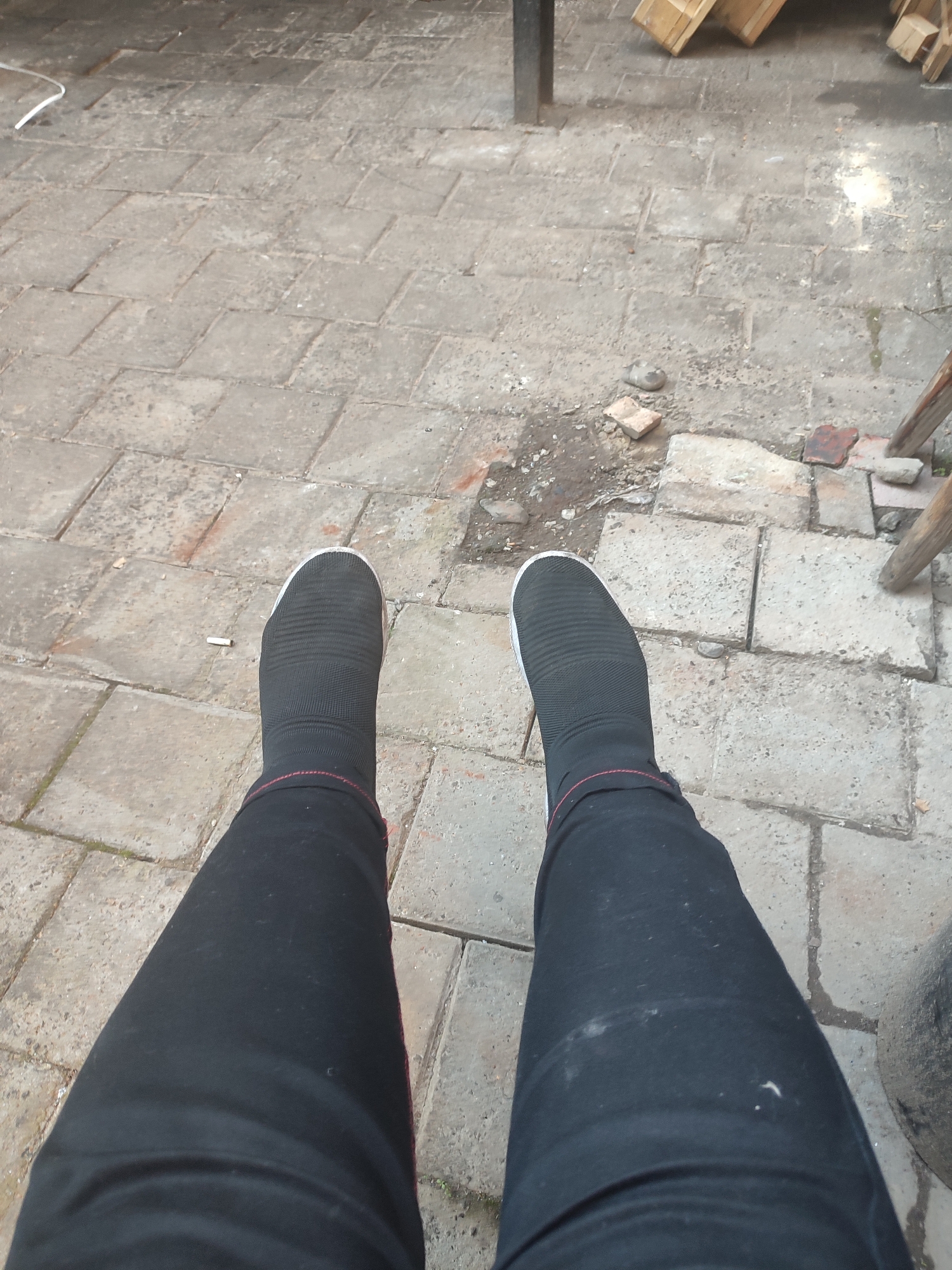 The image shows a close-up view of a person's legs wearing black socks and black pants, with black slip-on shoes. The person is sitting on a paved surface made of rectangular stone tiles. The environment appears to be outdoors, possibly a courtyard or walkway, with some debris and scattered building materials visible in the background. There is a cigarette butt on the pavement near the left foot. The overall scene is unremarkable and does not contain any specific visual indicators that would allow for pinpointing a location in Chernivtsi, Ukraine, or any other specific city.
