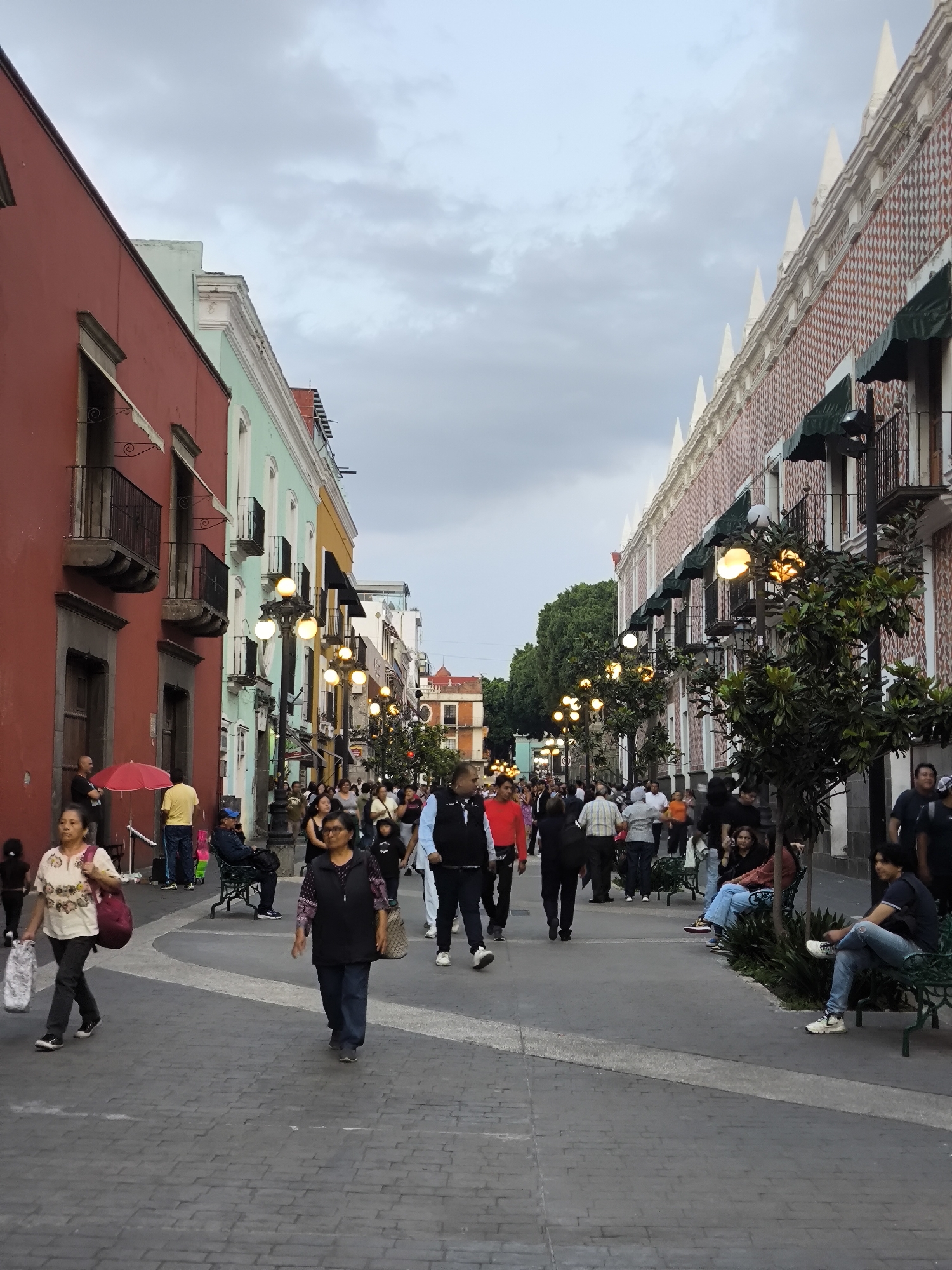 A street in Puebla, Mexico, is lined with colorful buildings and filled with pedestrians. The buildings on the left side of the street are painted red, turquoise, and yellow, and have balconies. The building on the right side of the street is decorated with red and white brickwork and has pointed white spires along the roofline. People are walking along the street and sitting on benches. Street lamps illuminate the scene. The sky is overcast.