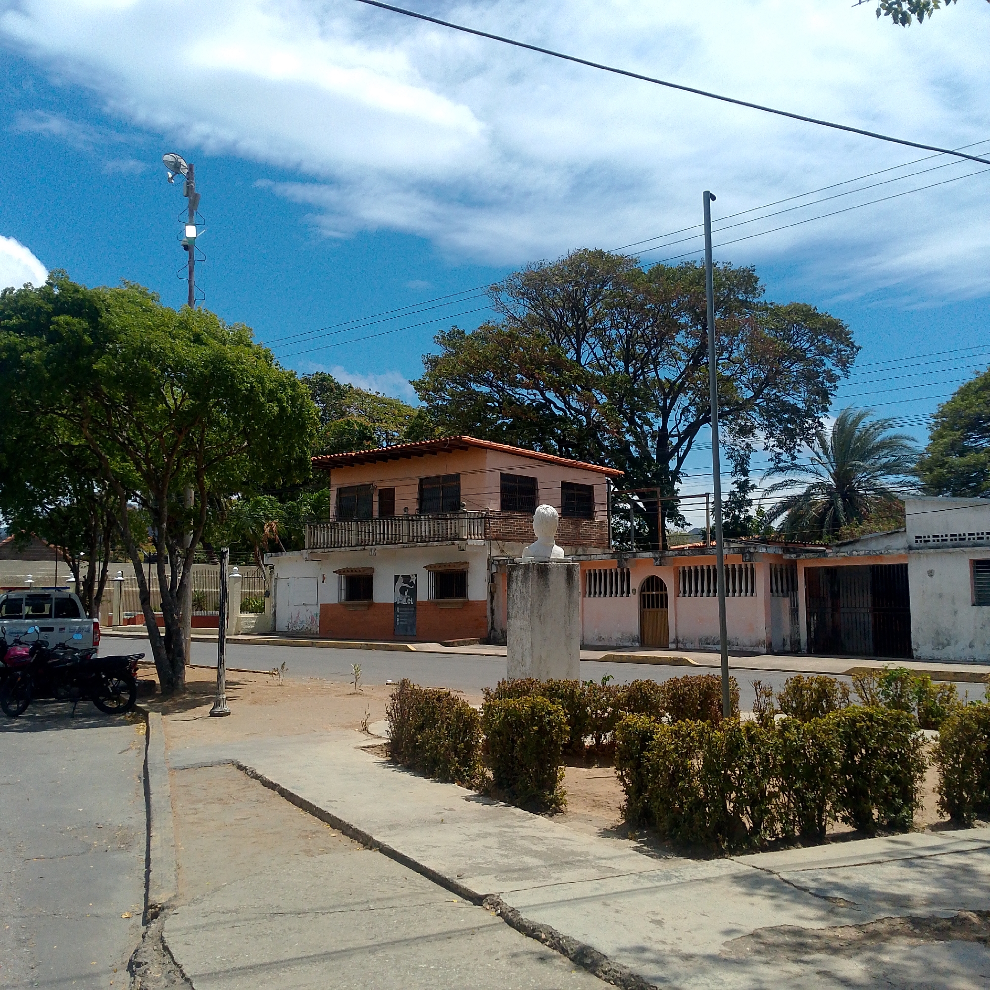 The image depicts a street scene in Altagracia, Venezuela. A two-story building with a peach-colored upper level and a white lower level with orange trim is visible. A bust on a concrete pedestal stands in front of the building. A row of neatly trimmed bushes separates the sidewalk from the street. A pickup truck and two motorcycles are parked near a tree to the left. Power lines crisscross the sky above. The sky is blue with scattered clouds. The ground appears dry and dusty. A street lamp is on the left.