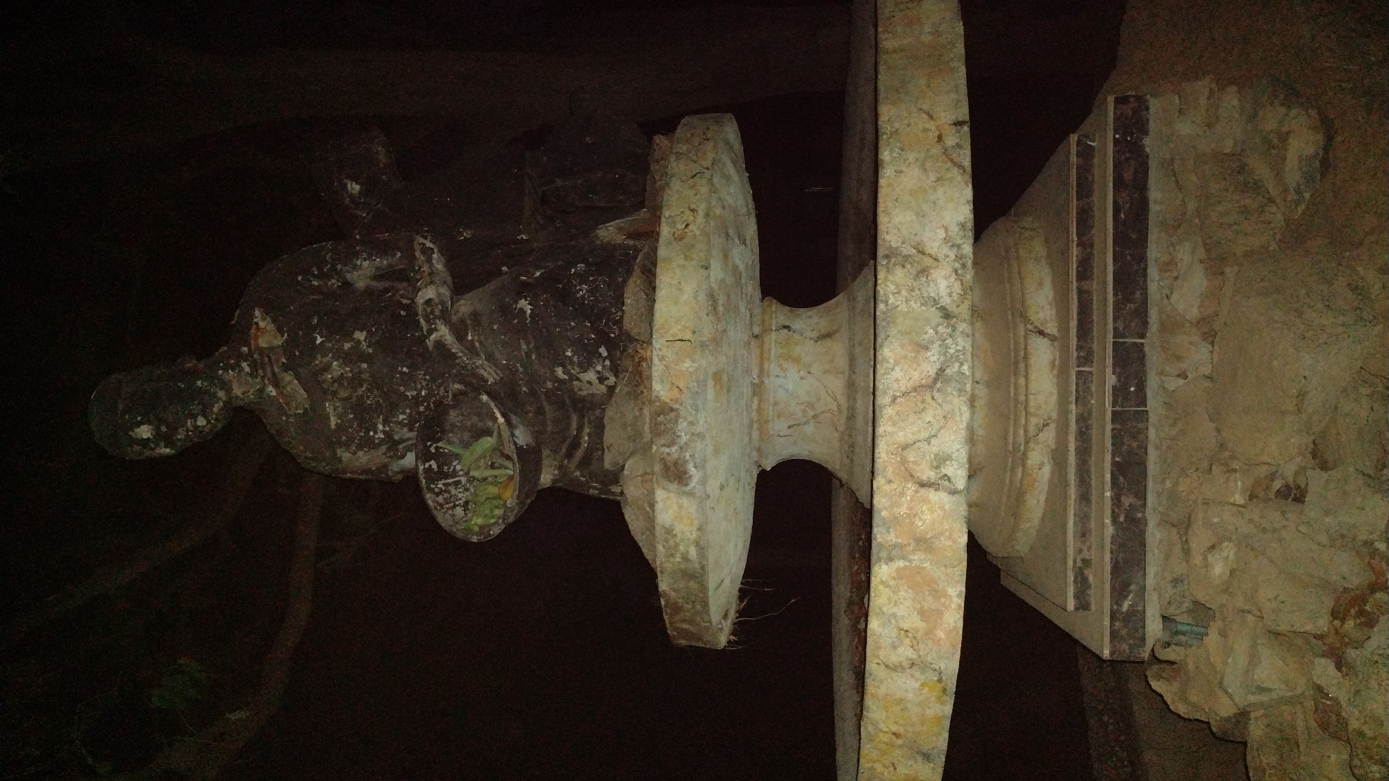 A statue, likely concrete or stone, is mounted on an elaborate, tiered plinth. The setting appears to be an outdoor or partially enclosed space, with a stone or earthen wall visible to the right. The lighting is poor, making details of the statue difficult to discern. The image's orientation is rotated counter-clockwise. The location is Jos, Nigeria.