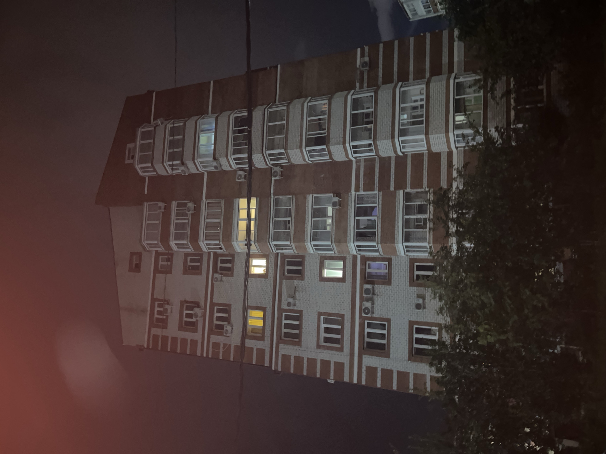 A low-angle, full-shot photograph taken at night shows the exterior of a multi-story apartment building in Chigiri, Russia. The building is predominantly constructed of red brick with white accents and features numerous windows, some of which are illuminated from within, indicating occupancy. The building has a unique architectural design with protruding sections that create a staggered facade. Air conditioning units are visible mounted below some of the windows. In the foreground, the tops of leafy green trees partially obscure the lower floors of the building. The sky above is dark, with a reddish glow appearing in the upper left quadrant of the image, possibly from artificial lighting or atmospheric conditions. Electrical wires are visible crossing the frame horizontally in the upper half of the image.