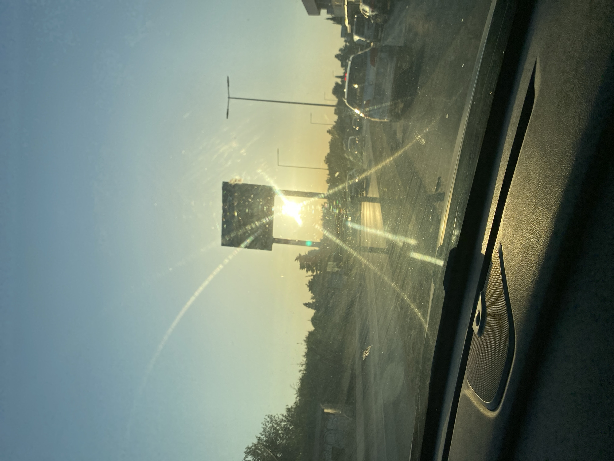 The image captures a sun-drenched scene from inside a vehicle, likely on a road in Bar, Montenegro. The sun, low in the sky, creates strong lens flare across the windshield, obscuring some details. In the middle ground, traffic is visible, with cars driving on a road that appears to be under construction or in disrepair, indicated by loose gravel and debris. A large signpost stands prominently, with the sun directly behind it. The background features a line of trees and vegetation, with a building visible on the right. The sky is clear and blue. The foreground shows the interior of the car, with the dashboard and a speaker grill visible.