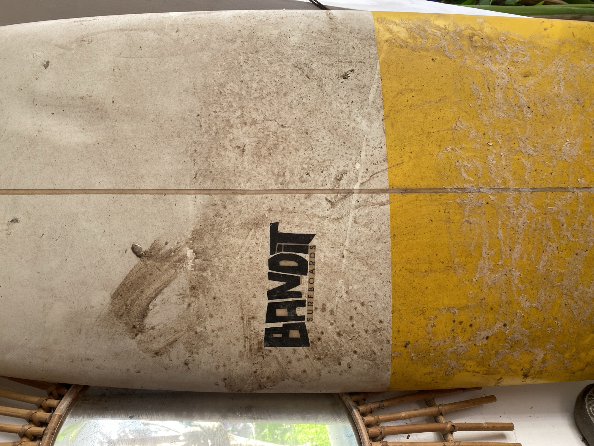A close-up, full-frame shot shows a dirty, weathered surfboard leaning against a white wall. The surfboard is split horizontally into two color sections: a dirty, off-white upper half and a bright yellow lower half. A black logo, reading "BANDIT SURFBOARDS," is centered on the upper half. The surfboard is covered in dirt, sand, and scuff marks. To the left of the surfboard, a portion of a rattan mirror frame is visible, with a reflection of greenery and what appears to be part of a building in the mirror. The setting is implied to be a storage area or a casual outdoor space in Kuta Selatan, Indonesia, potentially near a beach, given the surfboard and the sandy residue. No people or actions are depicted.