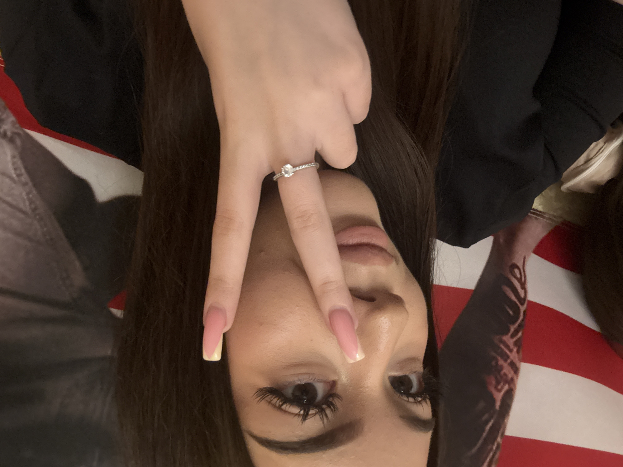 An eye-level, close-up portrait shows a person, likely female, posing for the camera. The image is rotated 90 degrees clockwise, presenting the person's head tilted to the left. The subject has long, dark hair framing their face.
The person's left hand is raised, making a V-sign or peace gesture, with the index and middle fingers extended upwards, partially covering their face. A silver-toned ring with a round, clear gemstone is prominently displayed on the index finger. The nails are long, shaped, and feature a French manicure style with a pink base and light, possibly yellow or cream, tips.
Visible facial features include expressive eyes with noticeable dark eyelashes, well-groomed eyebrows, part of the nose, and lips with a light-colored lip product. The person appears to be wearing a black top. On the person's right side, a patterned sleeve is visible, possibly a tattoo or part of their clothing, set against a background of red and white stripes consistent with an American flag. No text is visible. The overall mood is casual and posed.