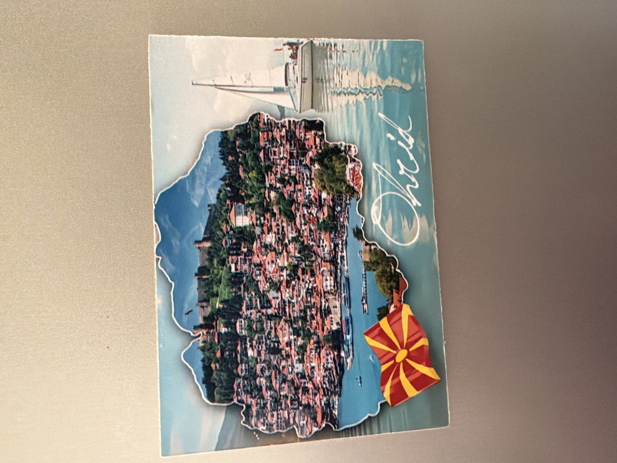 The image is a postcard featuring a scenic view of Ohrid, North Macedonia. The postcard depicts a silhouette of the city's historical architecture, including buildings with red roofs and a prominent castle on a hill overlooking a body of water. A sailboat is visible on the water to the right of the city. In the foreground, a Macedonian flag is affixed to the postcard. The word "Ohrid" is written in a cursive font on the water. The postcard is attached to a textured, neutral-colored surface, likely a refrigerator or a magnetic board, as evidenced by the lighting and the slight curvature of the postcard. There is no indication of the city of Pazardzhik, Bulgaria in this image.