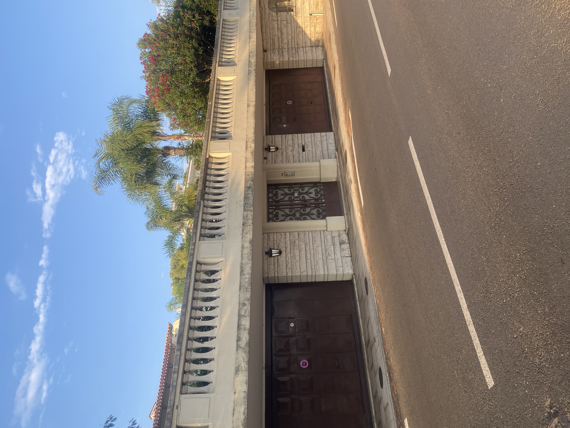 The image shows the facade of a building with a driveway on the left and two garage doors on the right, separated by a section of wall with a decorative metal door in the center. The building appears to be a residence, possibly a villa, located in Antibes, France. The facade is made of light-colored stone blocks and features a balustrade along the top. In front of the building is a paved area leading to the garage doors, and a street with white lane markings. Palm trees and flowering bushes are visible behind the balustrade, suggesting a sunny, Mediterranean climate. The sky is blue with some white clouds. The overall impression is one of a well-maintained, upscale property.