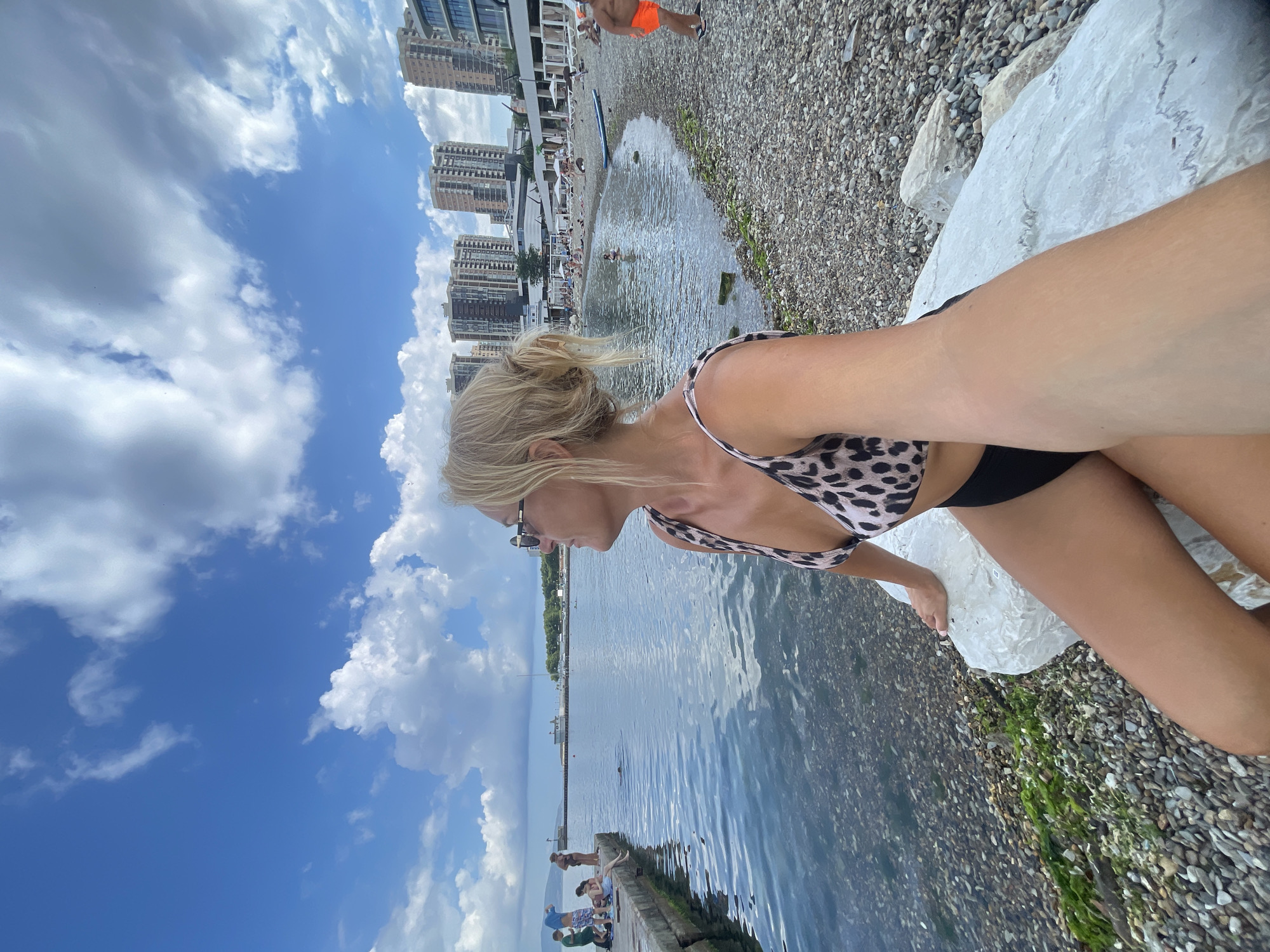 A woman with blonde hair tied up, wearing a leopard print bikini top and black bottoms, is sitting on a white rock on a pebble beach. She is looking out towards the sea, with buildings in the background. The sea is calm with some ripples. There are a few people visible on a pier in the distance, and more people on the beach further along. The sky is blue with scattered white clouds. The scene appears to be a resort area, likely in Novorossiysk, Russia, given the architecture of the buildings.