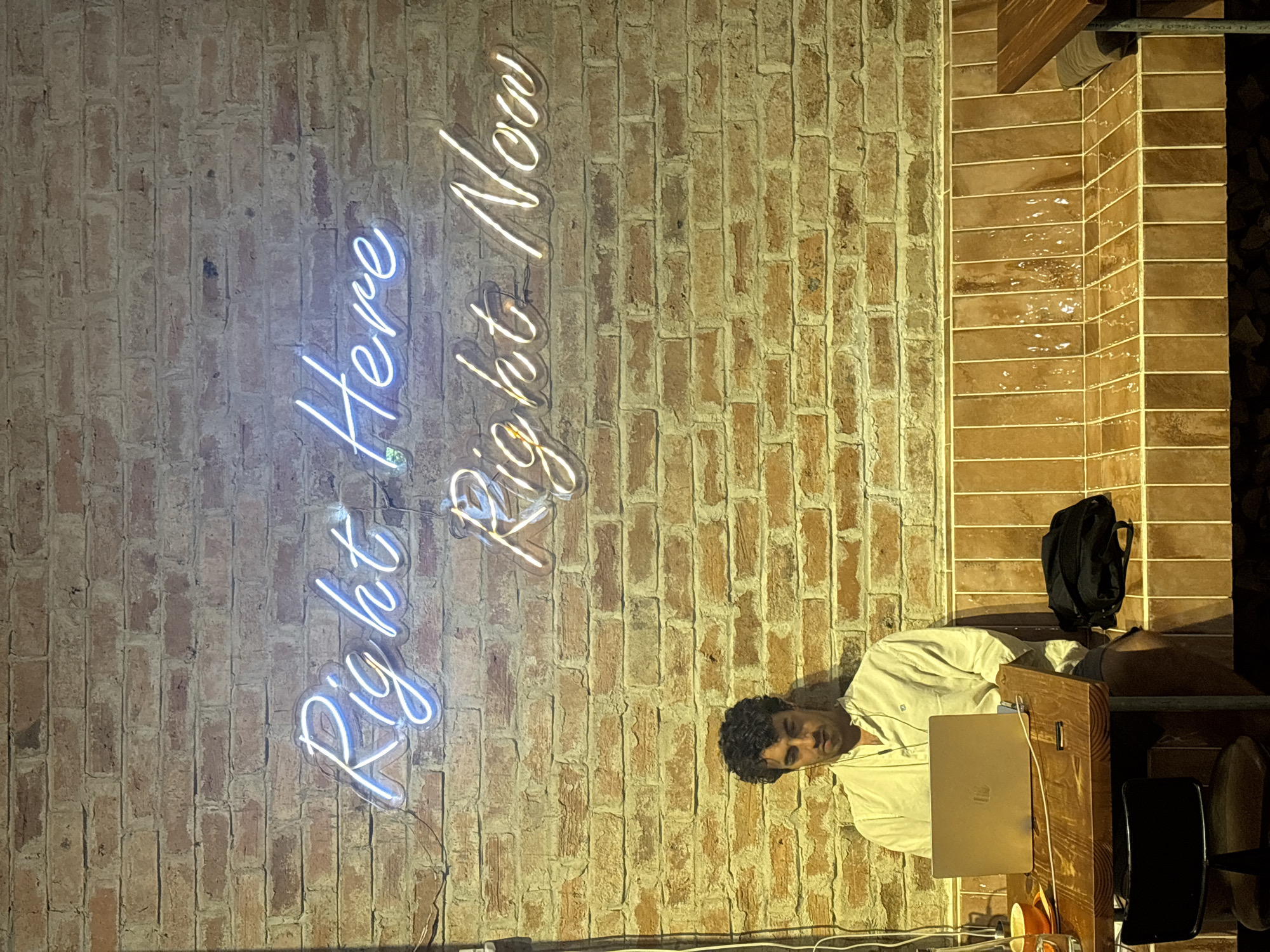 A man sits at a wooden table in front of a brick wall adorned with neon signs that read "Right Here Right Now". He is wearing a light-colored collared shirt and looking down at a laptop. A coffee cup sits on the table to his left, and a black backpack is on the bench behind him. The bench is tiled in a terracotta color. The scene appears to be indoors, likely a cafe or co-working space, in Da Nang, Vietnam.