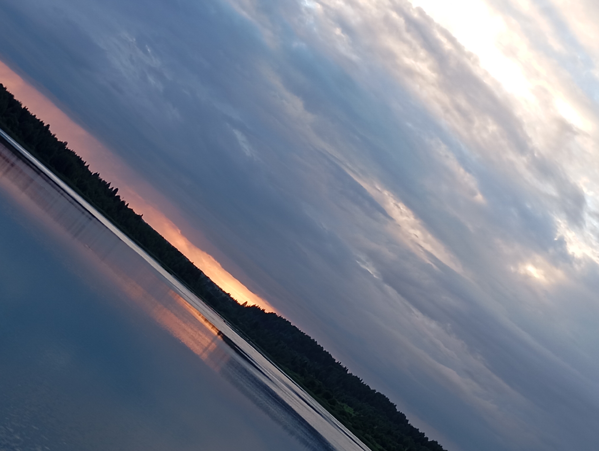 A diagonal shot captures a body of water in front of a forest line under a cloudy sky. The sun is setting or rising, casting an orange glow on the horizon, reflected in the water. The water surface is smooth with subtle ripples. The trees form a dark, dense line along the edge of the water. The sky is filled with dramatic clouds, ranging from dark gray to lighter shades, with streaks of sunlight breaking through. The overall scene suggests a tranquil, natural setting, possibly near the village of Ryabki, Russia.
