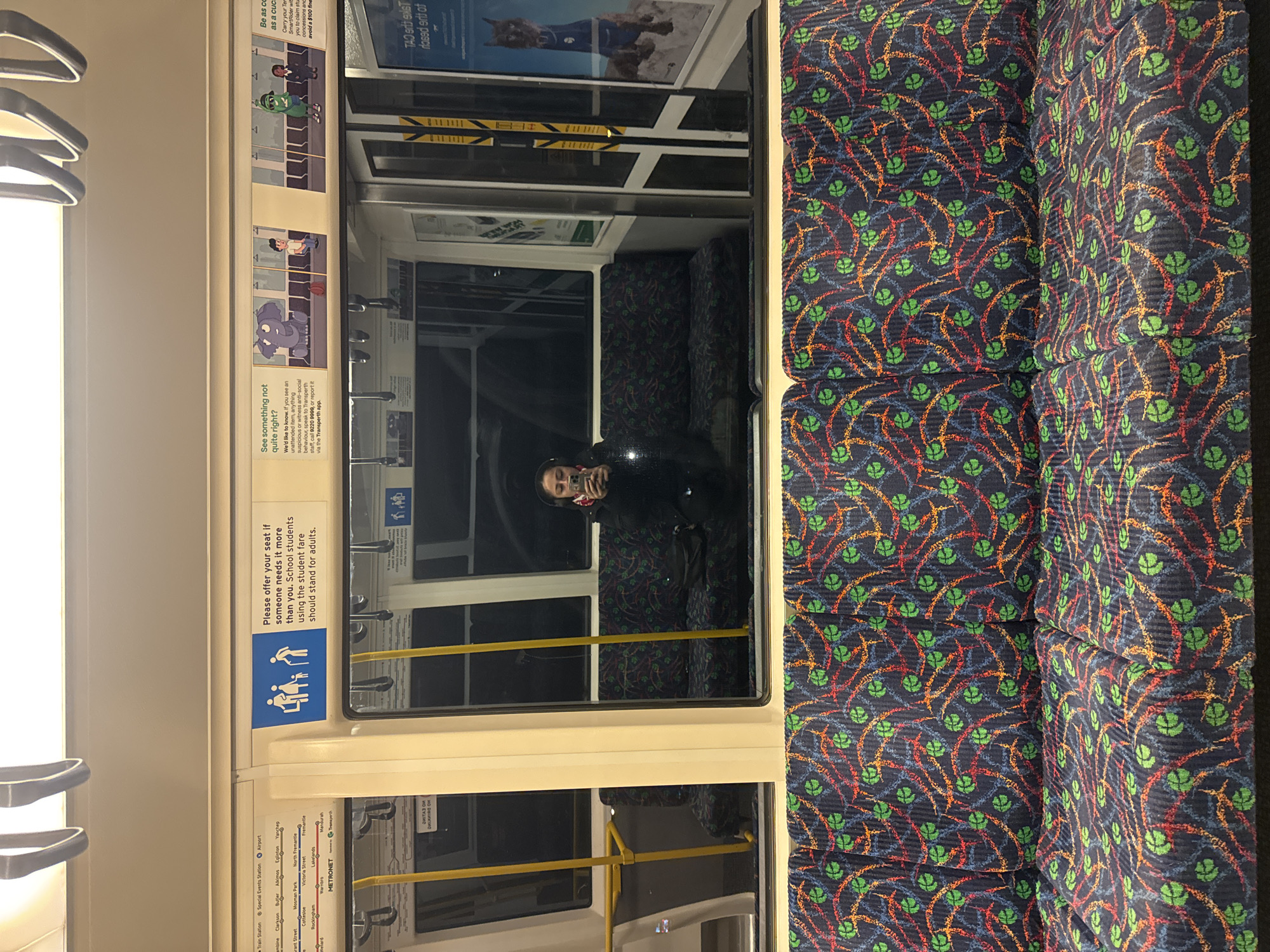 The image depicts the interior of a train carriage in Bassendean, Australia. The carriage has patterned navy blue seats with orange, red, blue, and green designs. A person is seated and is using their phone. Posters and signage are visible on the walls, including information about fare concessions and seat etiquette. The METRONET logo is displayed, indicating the train network.