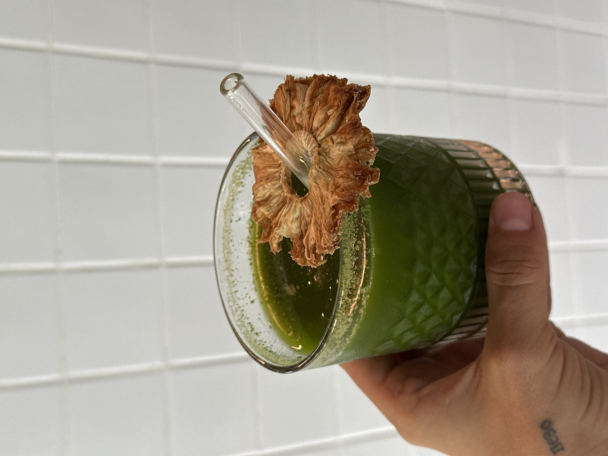 A person holds a patterned glass filled with a green smoothie, garnished with a dried pineapple ring and a clear straw. The background is a white, tiled wall. A tattoo is visible on the person's wrist. The location cannot be determined as Mengwi, Indonesia.