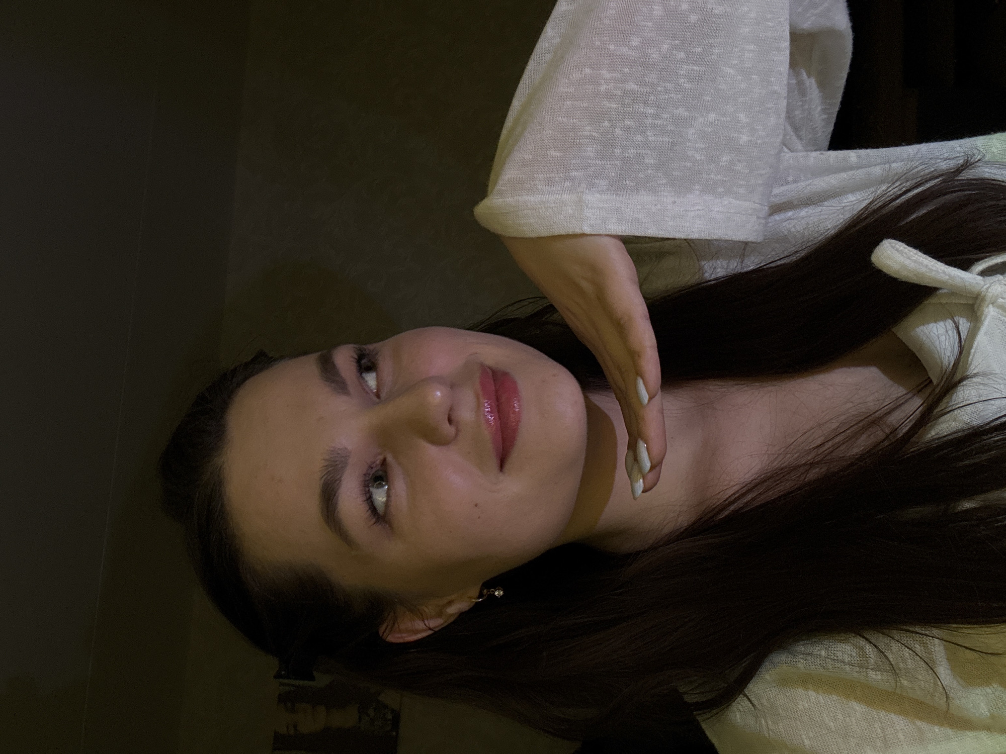The image features a young woman with fair skin, dark brown hair, and light-colored eyes, looking upwards and to her right with a soft, slightly smiling expression. Her lips are a glossy pink. She wears a light-colored, possibly off-white or cream, textured long-sleeved top. Her right hand (on the left side of the frame) is raised, with fingers gently touching her chin or jawline, revealing long, light-colored manicured nails. A small, dangling earring is visible on her left ear.
The subject is posed for a photograph, illuminated by a localized light source that highlights her features against a dark, indistinct background. This suggests an indoor setting with low ambient light, likely in the evening or at night. The mood conveyed is calm and contemplative. No discernible activity beyond posing is evident. There is no visible text in the image. The location context provided is Chaikovsky, Russia.