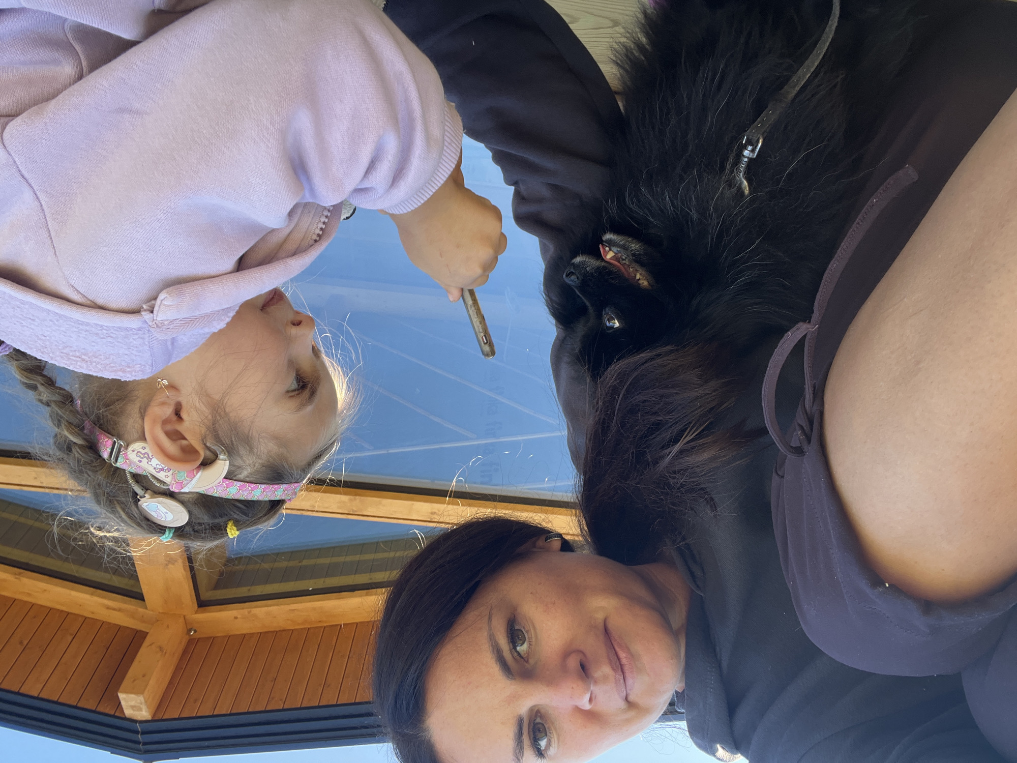 An adult woman and a young girl, both looking down, are outside. The woman has dark hair and is wearing a dark hoodie. She is holding a small, fluffy black dog in her arms. The girl has light brown hair in braids and is wearing a pink hooded jacket. She is holding a stick. A wooden structure with large windows is visible in the background. The sky is blue. The location is described as the city of Imeni Morozova, Russia.