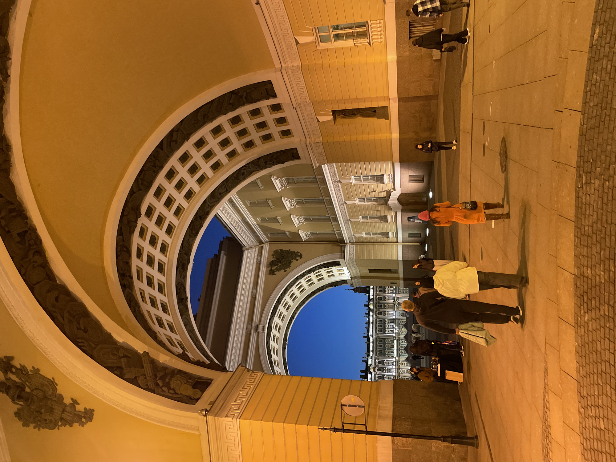 The image, correctly oriented, captures an evening scene featuring a grand architectural archway and several people in an urban setting. A monumental arch, constructed from light-colored stone or stucco, dominates the upper part of the frame, curving overhead. Its underside is elaborately decorated with square coffers and intricate bas-reliefs depicting classical figures or scenes. Warm artificial light from unseen sources illuminates the archway and the surrounding buildings, highlighting their architectural details.
Through the opening of the arch, a clear, deep blue night sky is visible, along with parts of other multi-story buildings in the distance, also illuminated from within their windows. The ground beneath the arch is a wide paved area, comprising large, light-colored flagstones that transition to cobblestones on the far right.
Several individuals are present on this paved area, suggesting casual movement. In the foreground, a person wearing a bright orange coat and a red knitted hat walks away from the viewer. Further back, another individual in a white jacket and dark pants stands, while a third person in a dark coat holds a light green object. Other figures, including one in a black coat and hat and another in a plaid shirt, are visible in the background. A lamppost with a circular sign stands on the right side of the paved area. The time of day is clearly night due to the dark sky and artificial lighting. No specific weather conditions are discernible beyond a clear sky. There is no visible text in the image.