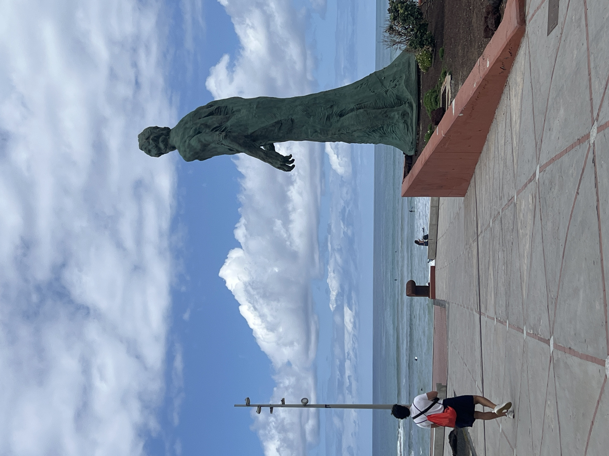 The image captures a scenic coastal view in Las Palmas De Gran Canaria, Spain, under a bright blue sky dotted with fluffy white clouds.
The primary subject is a tall, aged bronze-colored statue of a male figure, positioned on a raised, reddish-brown base or planter on the left side of the frame. The statue stands upright, appearing to wear draped clothing, and extends its arms slightly forward, gazing towards the ocean. Behind the planter, some sparse green vegetation grows.
To the right, a wide, paved promenade stretches along the coastline. The walkway features a light-colored tile pattern with visible intersecting reddish-brown lines. A person, seen from behind, walks along this promenade, moving away from the viewer towards the ocean. They are dressed in a white t-shirt, dark shorts, and white sneakers, carrying an orange or red backpack slung over one shoulder.
In the background, the expansive blue-green Atlantic Ocean is visible, appearing calm with subtle ripples. A few dark specks in the water might indicate distant individuals or objects. A slender utility pole with fixtures stands near the promenade, further in the background. The time of day is clearly daytime, characterized by bright, even lighting, suggesting late morning or early afternoon. The weather is clear and sunny with moderate cloud cover. No discernible text is visible within the image.