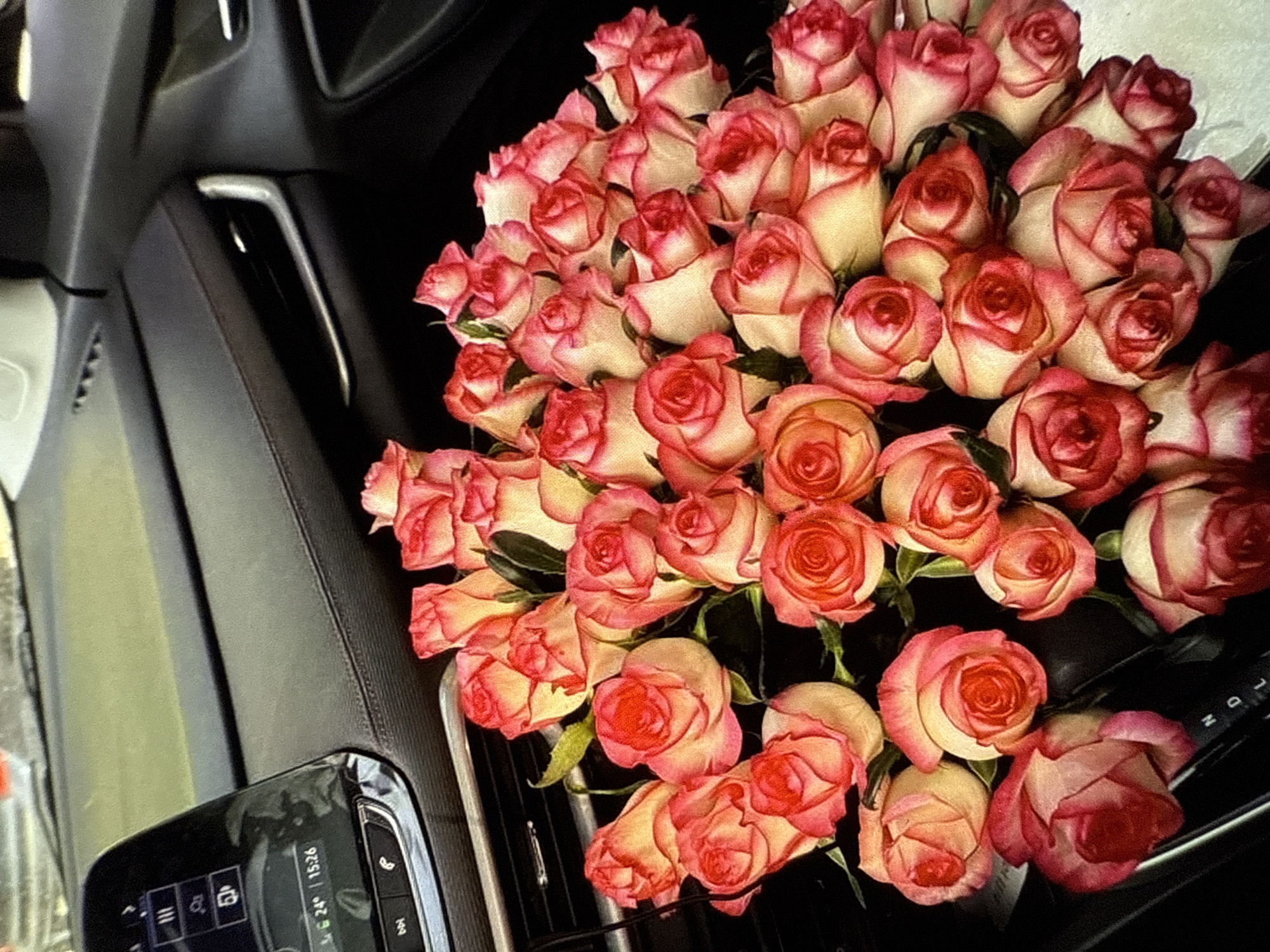 A bouquet of pink and white roses is placed in the passenger seat of a car, on top of the center console and gear shift. The dashboard and infotainment screen are visible in the background. The infotainment screen displays the current time as 15:26 and the temperature as 24 degrees. The gear shift indicates Park (P), Reverse (R), Neutral (N), and Drive (D). The lighting suggests it is daytime. There are no people visible in the image.
