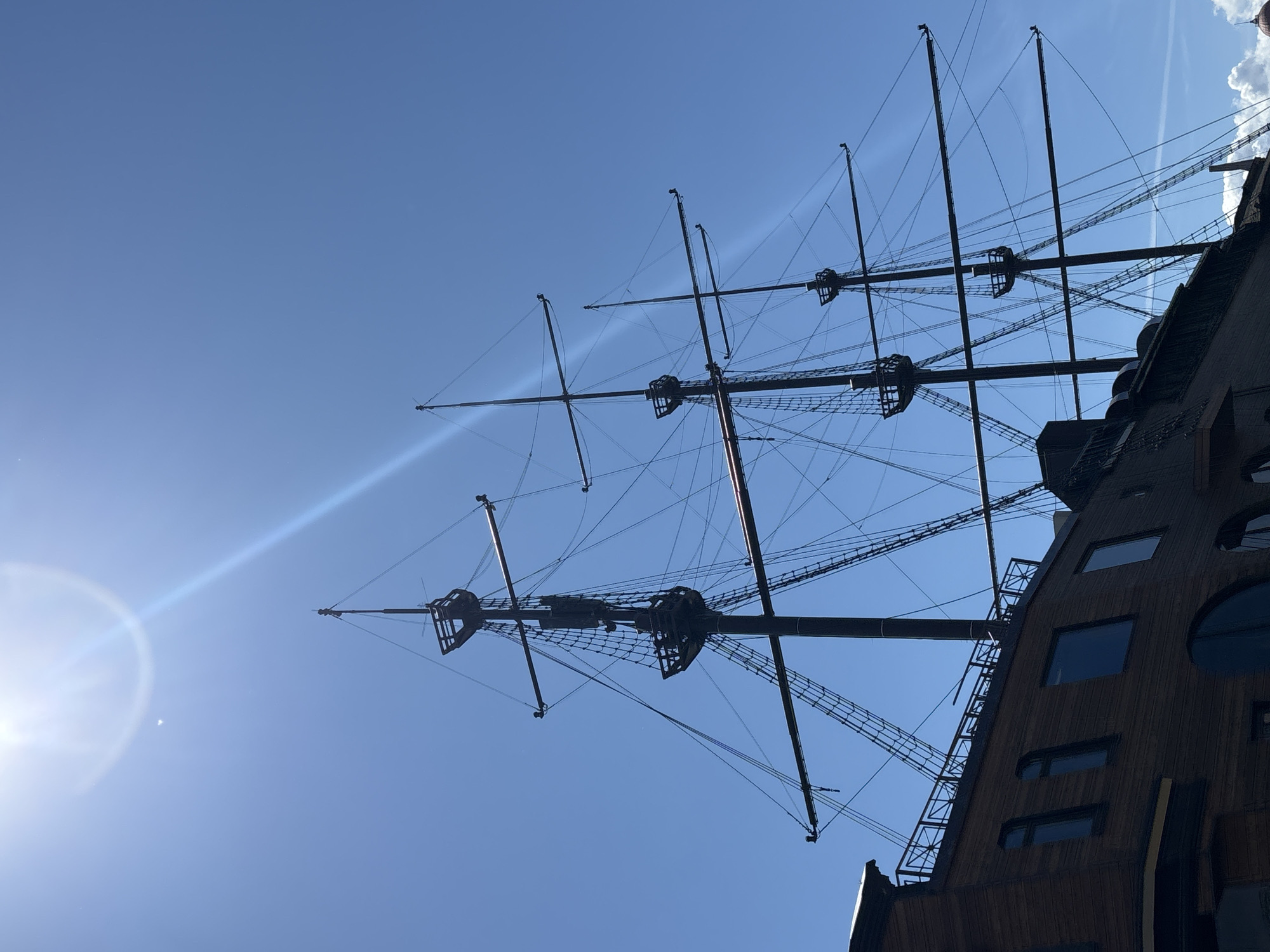 This is a low-angle, medium shot of a wooden sailing ship's masts and rigging against a bright blue sky with a sun flare in the upper left quadrant. The ship's wooden hull is visible on the left side of the frame, featuring several rectangular and arched windows. The masts are tall and dark, with multiple spars and an intricate network of ropes and rigging. Two crow's nests are visible on the main mast. The scene appears to be outdoors, with clear skies. This image was taken in Saint Petersburg, Russia, likely near the Neva River or the Gulf of Finland, where such replica ships are often found as tourist attractions or restaurants. There are no people visible in the image.