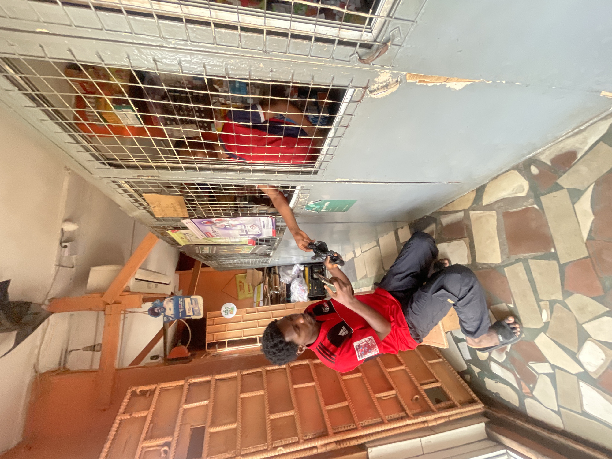 A man in a red shirt and dark pants is kneeling on a tiled floor, holding a smartphone in his left hand and a black object in his right hand. He is looking down at his hands. Another person's arm is visible through a barred window, holding a similar black object. The scene appears to be a storefront with a barred window and shelves stocked with goods behind the window. The environment suggests a commercial setting in Akowonjo, Nigeria.