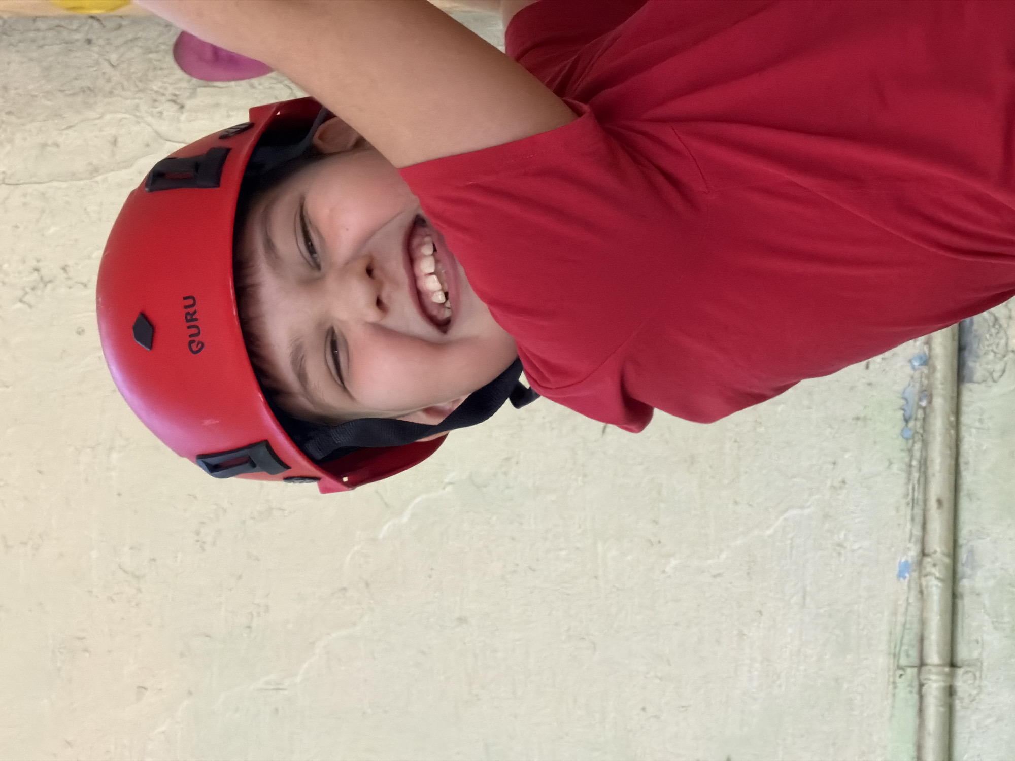 A child wearing a red "GURU" branded helmet and a red t-shirt is smiling upside down. The background appears to be an indoor wall with some discoloration and a pipe running vertically. The exact location in Izhevsk, Russia, cannot be determined from the provided image.