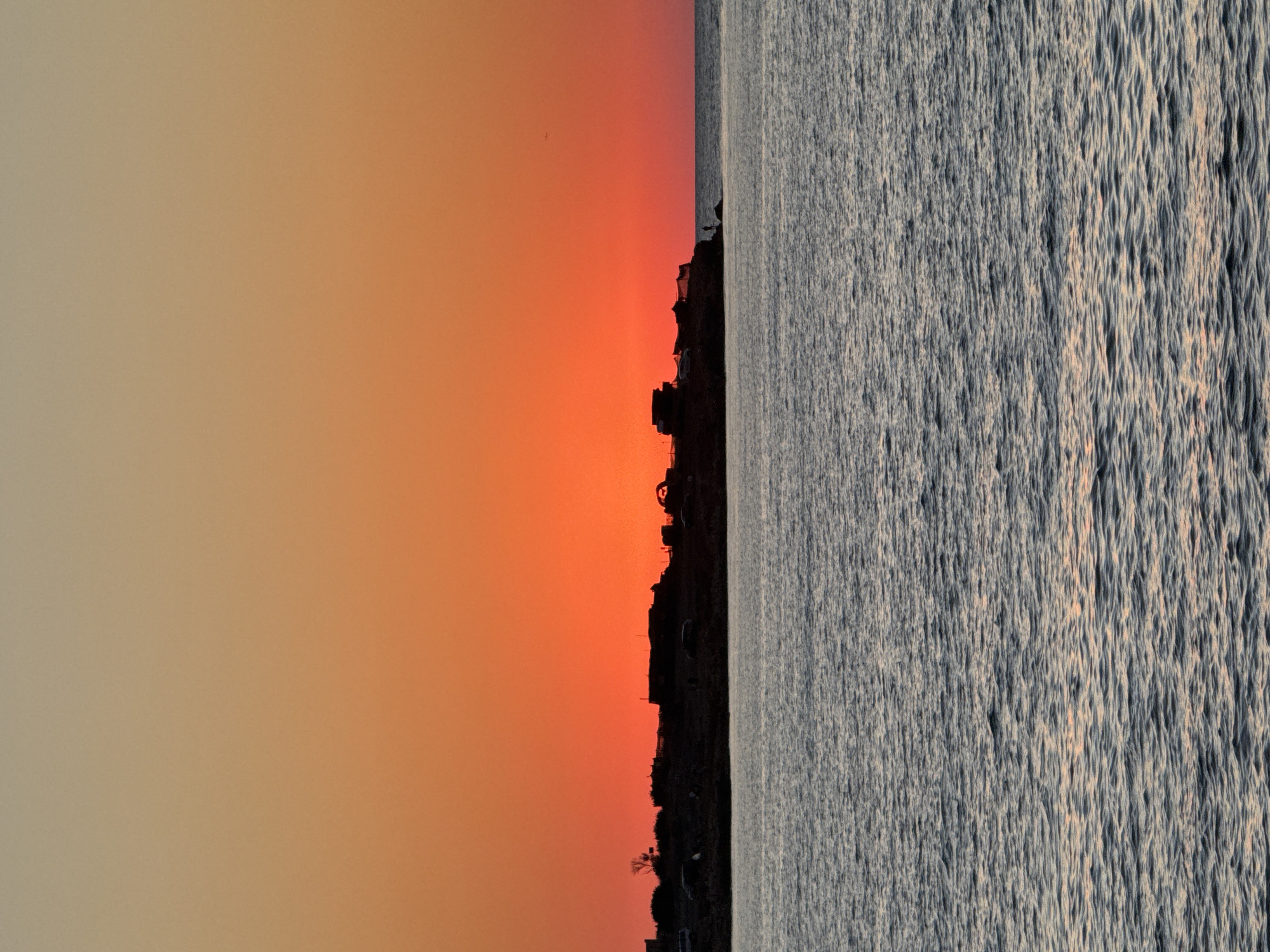 A silhouetted landscape at sunset depicts a body of water with a distant shoreline populated by dark, indistinct structures and vehicles. The sky transitions from a vibrant orange near the horizon to a lighter peach hue above. The water is rippled, reflecting subtle hints of the sunset's colors. A solitary figure stands on the right edge of the shoreline. The scene is characterized by strong backlighting, creating a dramatic silhouette effect against the bright sky. It is not possible to determine the specific location in Sevastopol, Ukraine from the provided image.