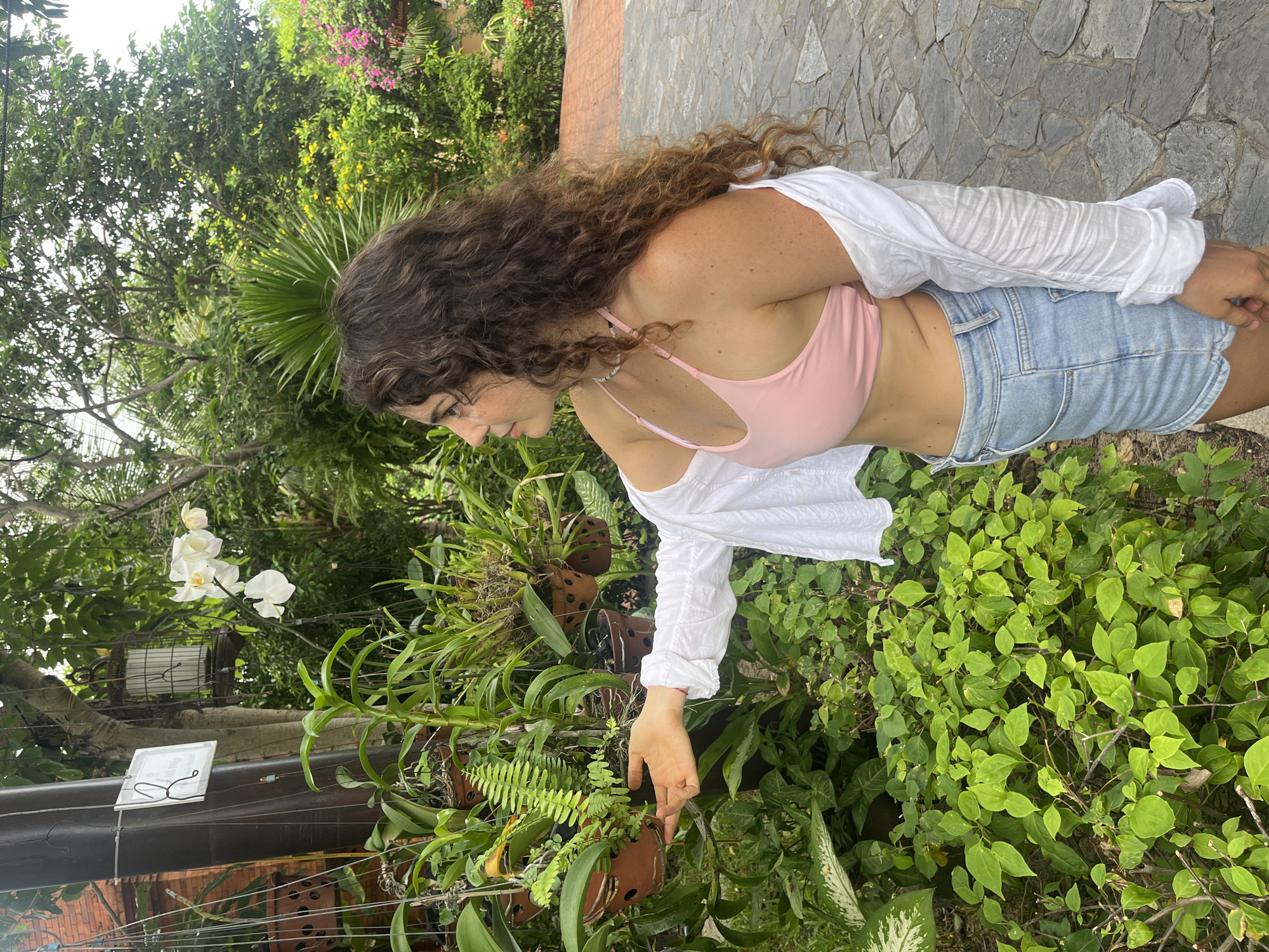 A young woman with long, curly brown hair is standing in a garden in Phu Thuy Ward, Vietnam. She is wearing a pink bikini top, denim shorts, and a white open shirt. Her left hand is extended, touching a plant. The garden features various potted plants, including ferns and orchids, hanging from a wooden pole and overhead structures. Lush greenery and trees surround the woman, and a stone pathway is visible to her right. The lighting suggests it is daytime.