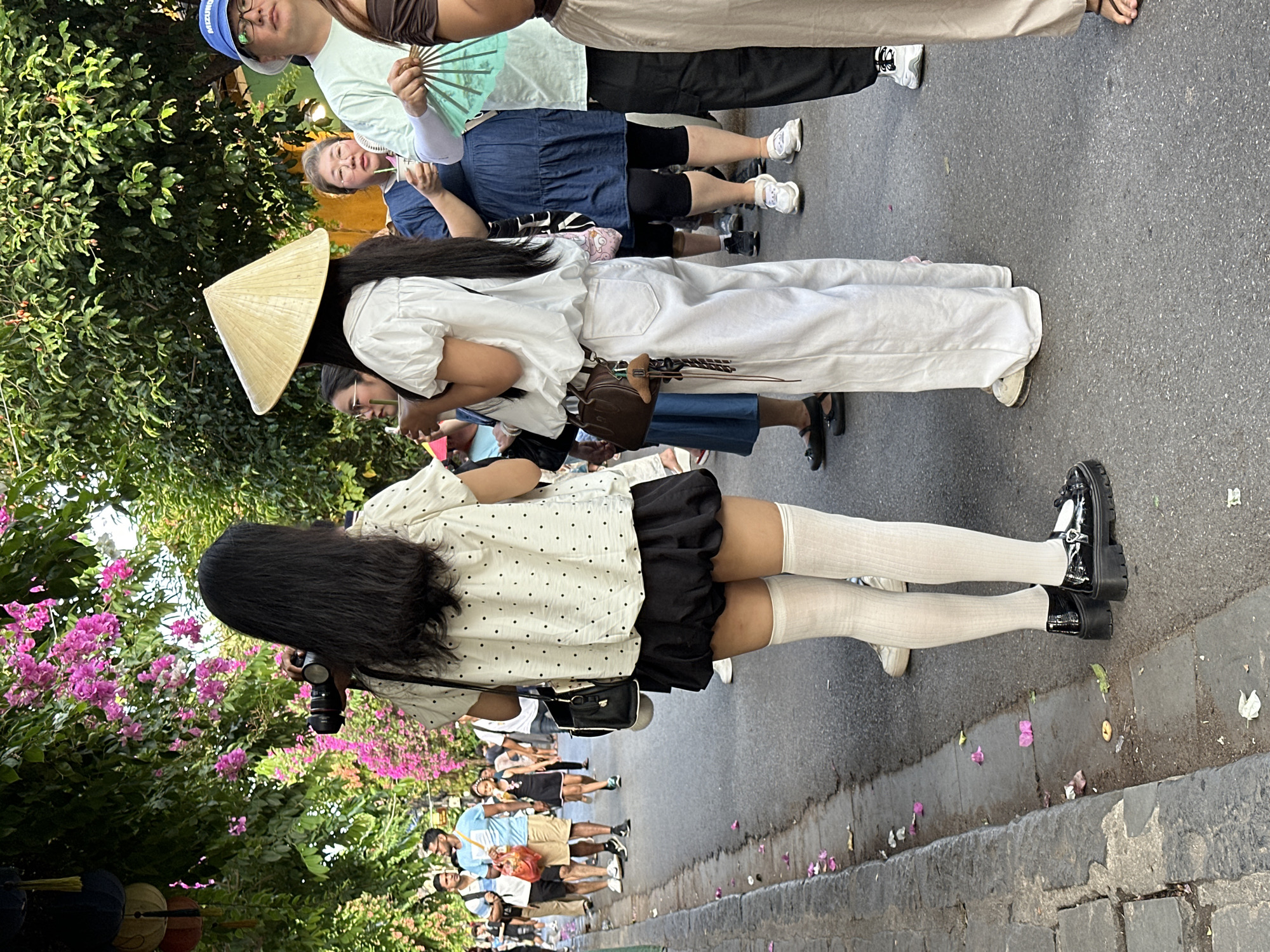 In Hội An Ward, Vietnam, on a paved street lined with trees and hanging lanterns, two young women are the central focus. The woman on the left, with long black hair, wears a white polka-dot blouse and a black bubble skirt, complemented by white knee-high socks and black platform shoes. She holds a camera. To her right, a woman wearing a conical hat is dressed in a white puff-sleeved shirt and loose white pants. A small brown purse is slung across her body. Several other people are visible in the background, some holding fans, and pedestrians are walking along the street, suggesting a busy, possibly tourist-oriented area. The ground is littered with fallen pink petals and debris.