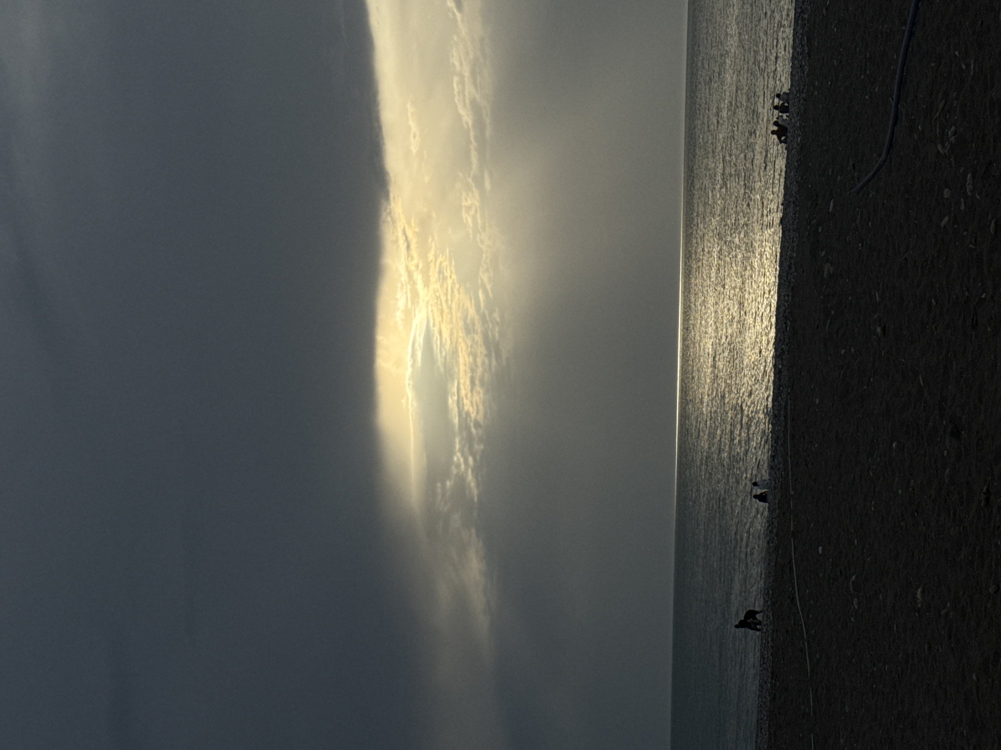 The image captures a view of the Black Sea coastline in Batumi, Georgia. On the right, a dark pebbly beach is visible, with a few figures appearing to walk along the shore. A single thin white rope lies along the beach. The sea reflects sunlight, creating a bright streak across the water's surface. The sky is dominated by heavy, dark clouds on the left, contrasting with a brighter, sunlit area toward the center.