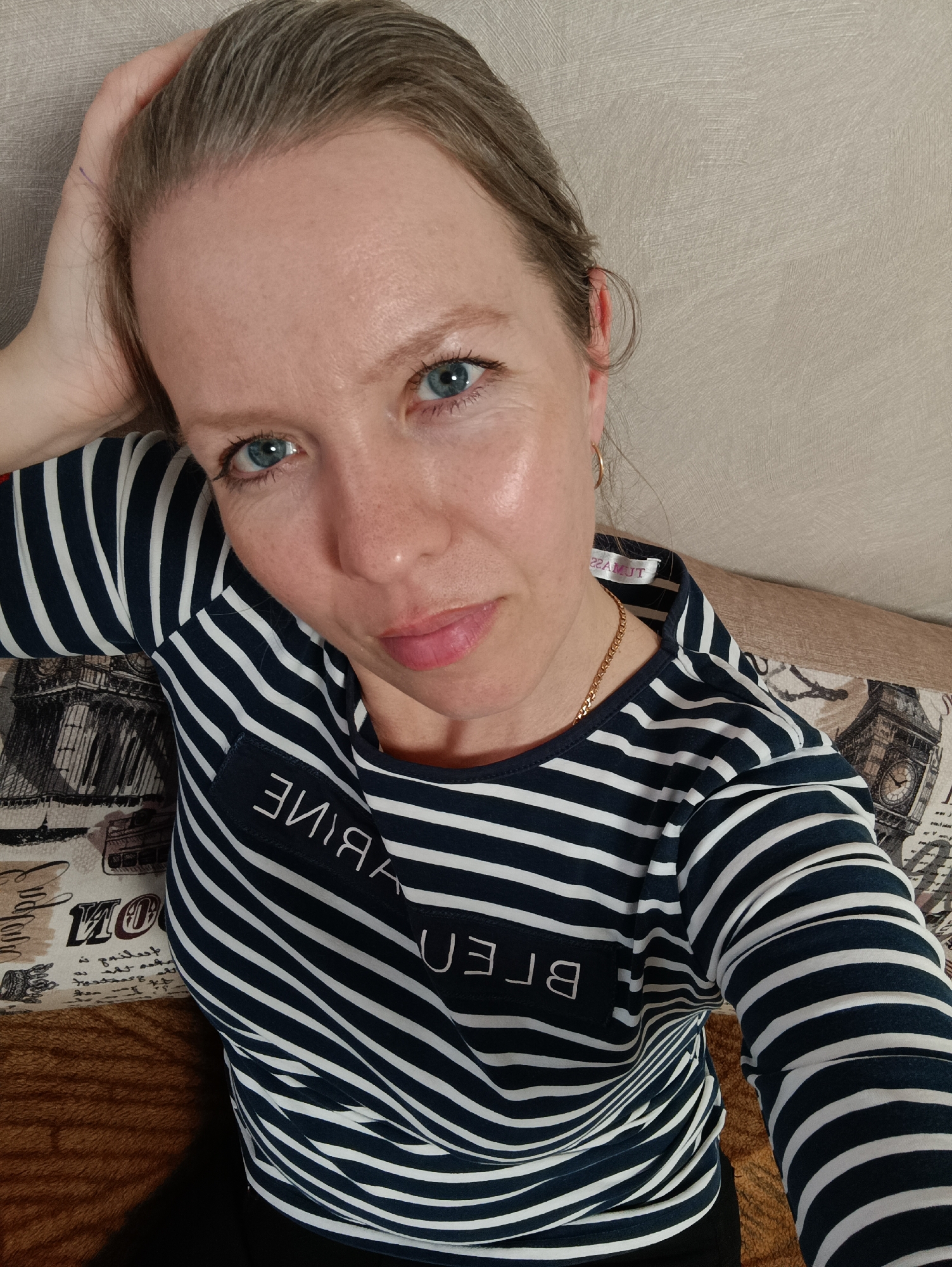 The image captures a close-up, eye-level indoor selfie of a woman taken in Chaikovsky, Russia. She has light skin, prominent blue eyes, and light brown or blonde hair styled loosely. She looks directly at the camera with a neutral to slightly thoughtful expression.
She is wearing a dark blue and white striped long-sleeved shirt. Text printed on the shirt's front, which appears reversed in the selfie, reads "BLUE" and "MARIN." A thin gold chain necklace is visible around her neck, and a small gold hoop earring can be seen in her right ear. Her left hand is resting gently on the top of her head, touching her hair.
The woman is seated or reclining, with a patterned fabric covering a surface behind her, likely a couch or bed. This fabric features sketches of cityscapes and landmarks, including Big Ben and the word "London," alongside other illegible text. Adjacent to this is a section of brown, textured fabric. The background wall is a light, neutral tone with a subtle, textured finish. A shirt label, also reversed, showing "TUMASS" is visible inside her collar. The lighting is soft and consistent with an indoor setting.