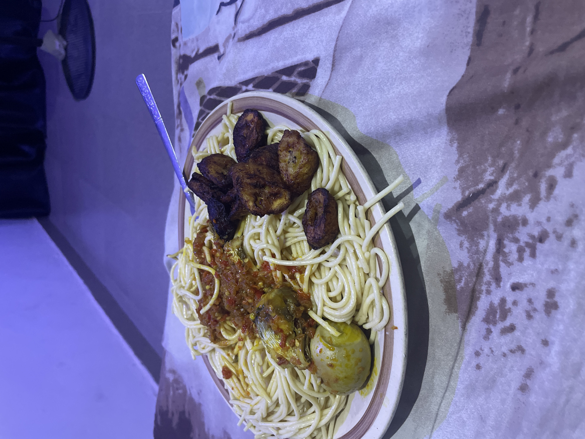 A plate of spaghetti is served with fried plantains, a hard-boiled egg, and a fish head in a tomato-based sauce. The plate sits on a patterned cloth, possibly a bedsheet, with a textured surface. The lighting suggests an indoor setting, possibly at night, with a purple hue cast over the scene. The background shows a wall and a dark area that might be furniture. The overall impression is of a simple, home-cooked meal. The location is identified as Barako, Nigeria.