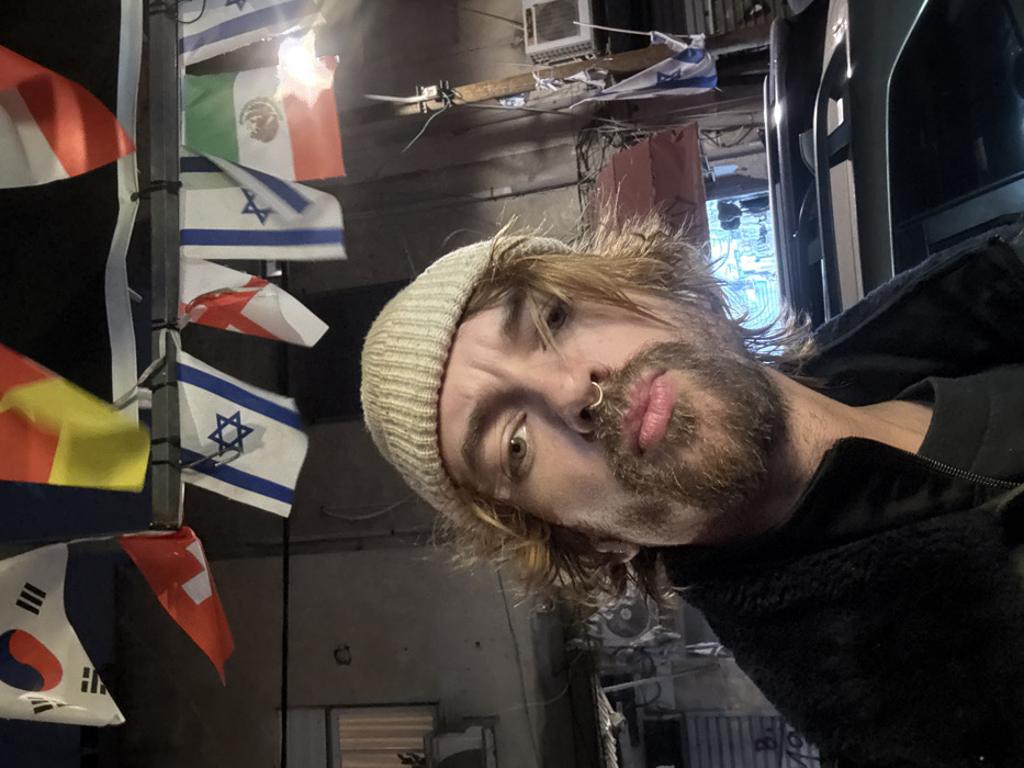 A selfie features a person with shoulder-length blond hair, a beard, and a nose ring. They wear a light-colored beanie and a dark jacket. The camera angle is slightly tilted. In the background, a pole displays flags from different nations, including South Korea, Israel, and Mexico. The setting appears to be outdoors in a somewhat cluttered area with visible wiring and building fixtures. The lighting is dim.