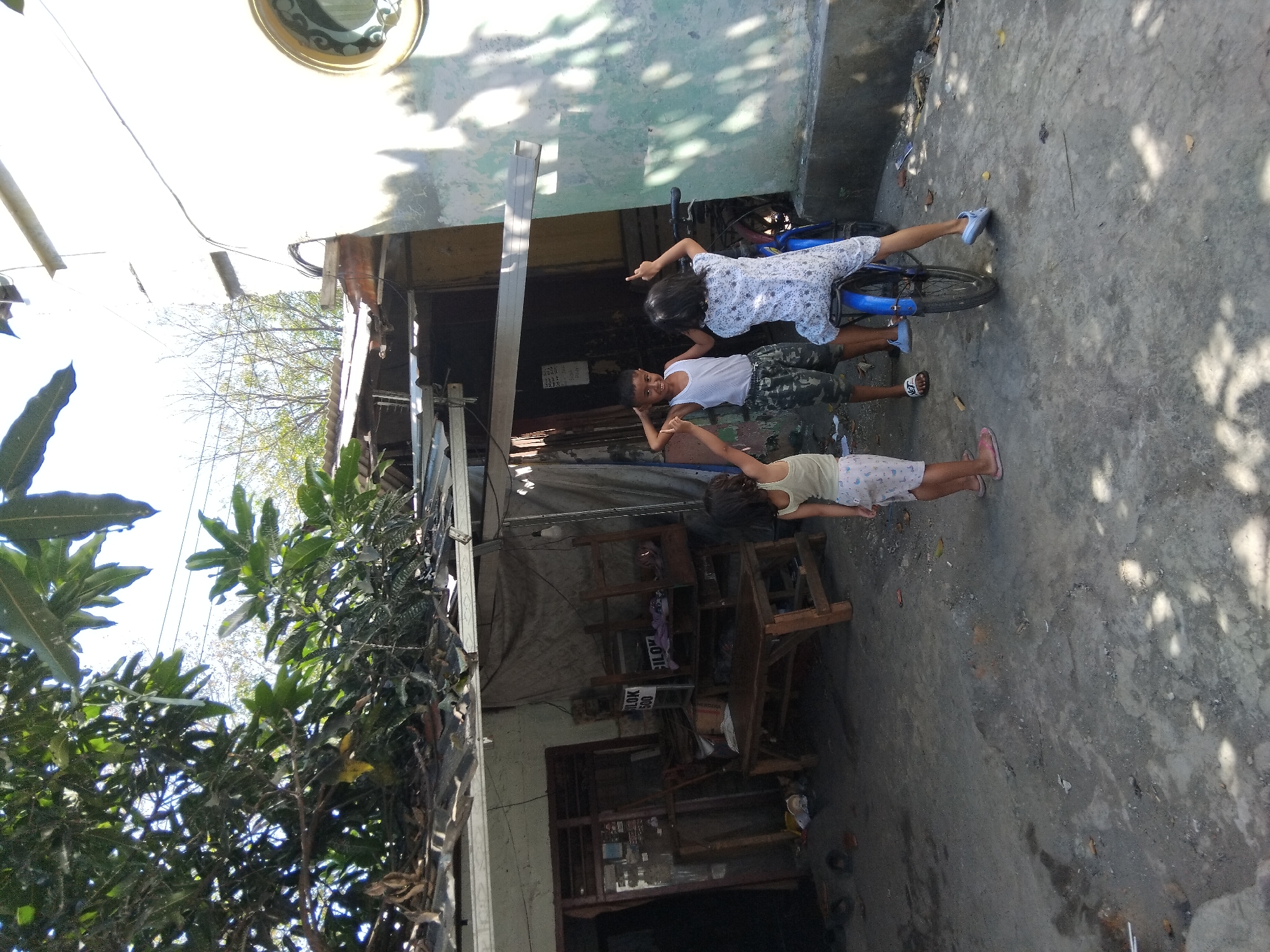 In an outdoor setting with a concrete ground and shadows from overhead foliage, three children are interacting near a dilapidated structure. On the left, a young girl in a light-colored top and patterned shorts stands facing away from the camera, her right arm slightly raised. In the center, a young boy wearing a white tank top and camouflage shorts stands with his hands on his head, looking towards the camera. To the right, another child, appearing to be a girl, is seated on a blue bicycle, facing away from the camera and raising her right arm. The background includes a structure with a corrugated metal roof, tarpaulin sheeting, wooden furniture, and a white wall with a circular window. The overall scene suggests a casual, everyday moment in a residential area, possibly in Berbek, Indonesia, given the architectural style and vegetation.