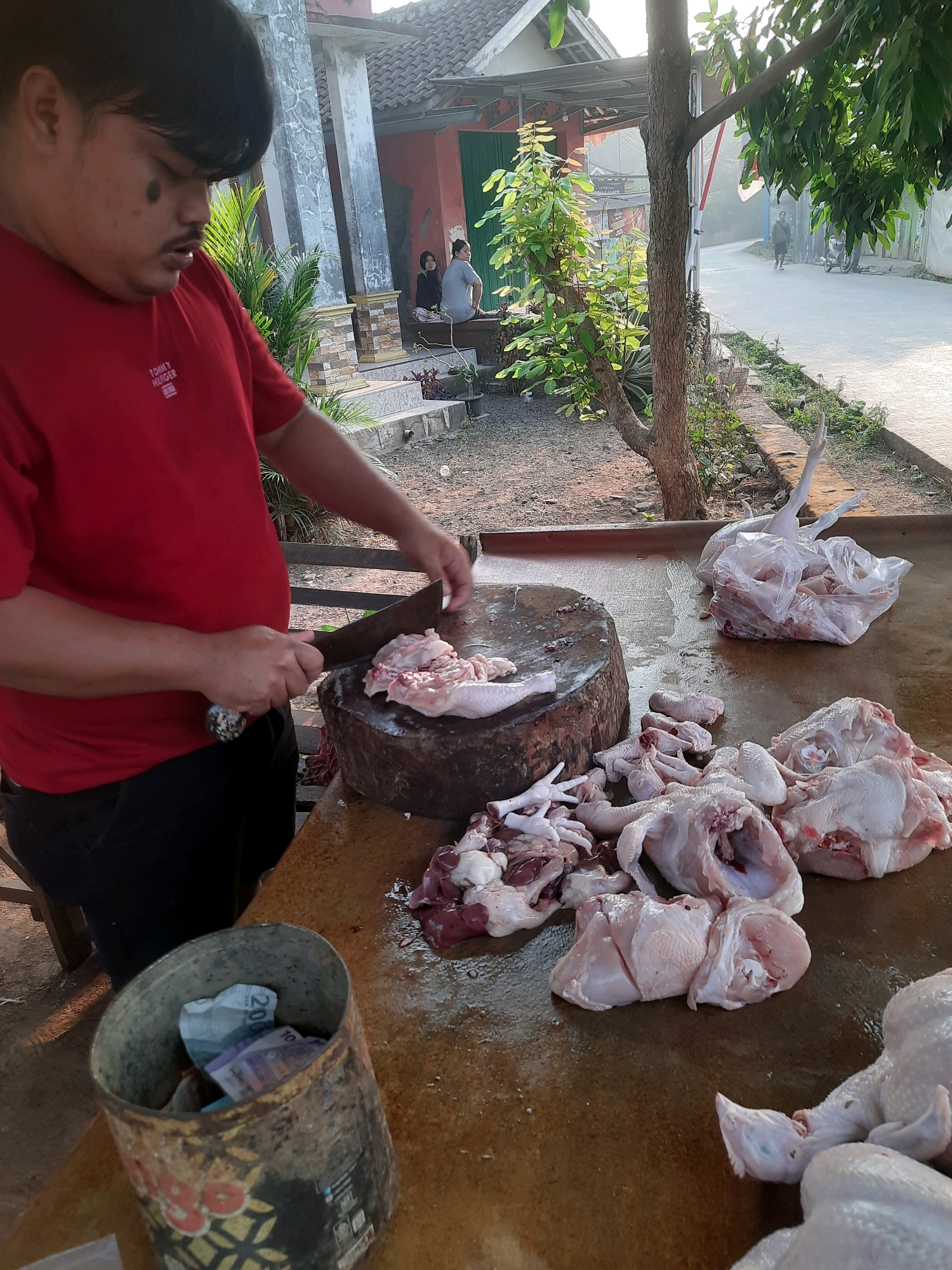 The image depicts a man in a red t-shirt and black pants in Gandoang, Indonesia, standing at a wooden table, actively butchering chickens. He is using a large cleaver to cut raw chicken parts on a wooden chopping block. The table surface is wet and appears to be made of wood, with numerous pieces of raw chicken scattered across it, including whole chickens, parts, and organs. To the left of the butcher, a metal container is visible, containing folded banknotes. In the background, a building with a red facade and white pillars is present, along with some trees and a road. Sunlight indicates it is daytime.