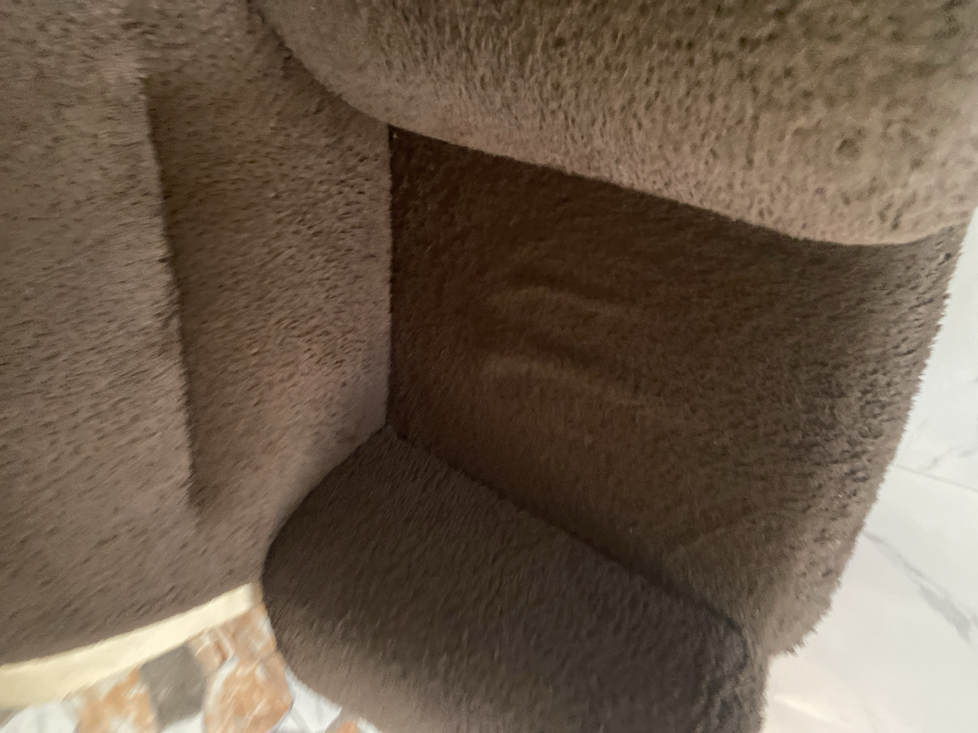 This media file shows a close-up view of the interior of a brown, plush pet bed. The fabric is soft and textured, with a raised nap. The bed appears to be constructed with padded sections, creating corners and enclosed spaces. In the lower left corner, a sliver of a cream-colored edge is visible, suggesting the outer rim or base of the bed. Below this edge, a portion of tiled flooring can be seen, with tiles featuring earthy tones of brown, orange, and cream, arranged in a mosaic or irregular pattern. The lighting is somewhat dim, creating shadows within the bed, and the overall impression is one of a cozy, enclosed space. No people, animals, text, or specific time of day or weather cues are visible. The location context provided is Jauro Yinu, Nigeria, but this information is not discernible from the image itself.