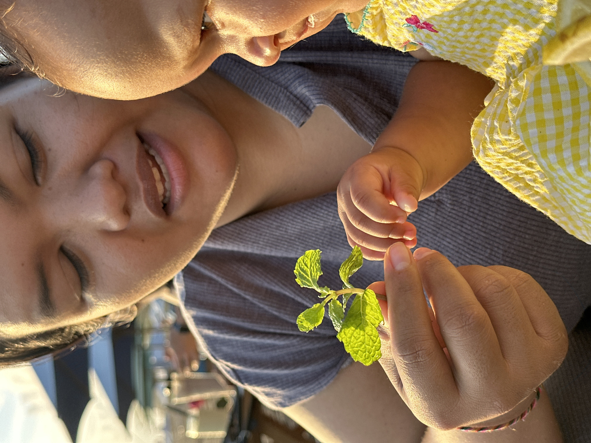 A close-up shot shows a woman with closed eyes, smiling slightly, being kissed on the forehead by a baby wearing a yellow and white checkered garment. The woman is wearing a grey shirt and is holding a sprig of green leaves with one hand while the baby's hand is near the sprig. The background shows blurry outdoor elements. The location is possibly North Kuta, Indonesia, based on potential contextual cues.