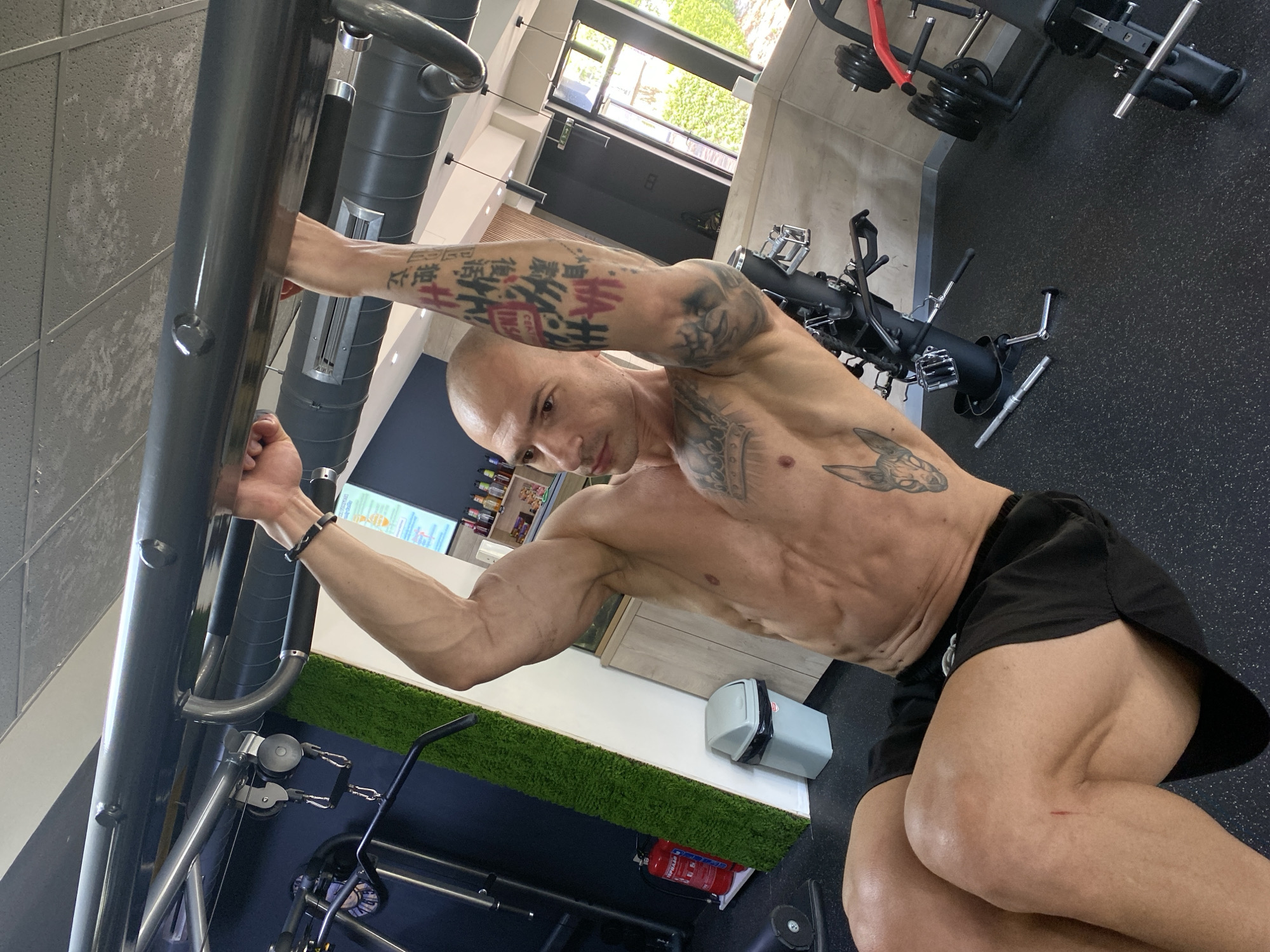 A muscular, bald man is the central subject of this image, captured in an indoor gym, likely in Pleven, Bulgaria. He is shirtless, wearing black athletic shorts, showcasing a very defined physique with prominent abdominal muscles and visible veins in his arms and legs. He looks directly at the camera with a neutral expression.
The man is positioned in the foreground, leaning against or holding onto a large, dark grey piece of gym equipment, possibly a pull-up station or part of a cable machine system. His left hand is gripping a thick bar, while his right arm is extended. Both of his arms and his torso are heavily tattooed with intricate designs, including what appears to be East Asian script on his left forearm, some characters highlighted in red. A black bracelet is visible on his left wrist. A small red scrape or cut is present on his right thigh.
The gym features black rubber matting on the floor and a section with artificial green turf running along a wall. Various other pieces of gym equipment, such as weight benches, weight plates, and components of cable machines, are discernible in the background. Large windows allow natural light to illuminate the space, revealing green foliage outside, indicating it is daytime. Overhead, exposed ventilation ducts and modern lighting fixtures are visible against a textured ceiling. Other objects include a grey trash can with a black liner and a red fire extinguisher on the floor near the turf area. The number "8" is visible on a door frame through one of the windows.
