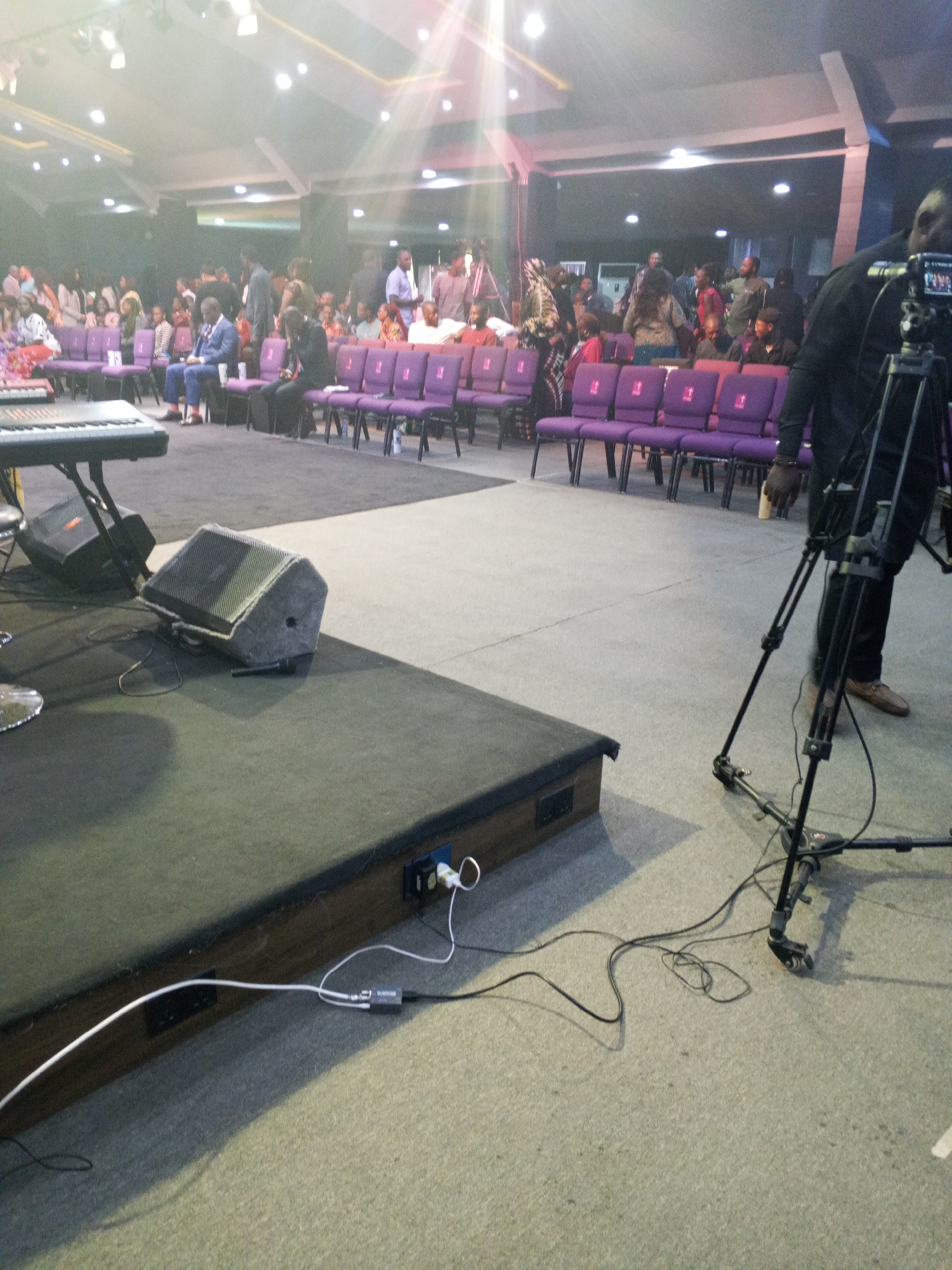 The image shows an indoor scene in a likely church setting in Abuja, Nigeria. A stage covered in black carpet is in the foreground, with audio equipment, including a speaker and keyboard, visible. Electrical cables run from the stage. In the background, rows of purple chairs are occupied by numerous people. A camera on a tripod is positioned to the right, with a person standing behind it. The lighting is bright, possibly due to stage lighting.