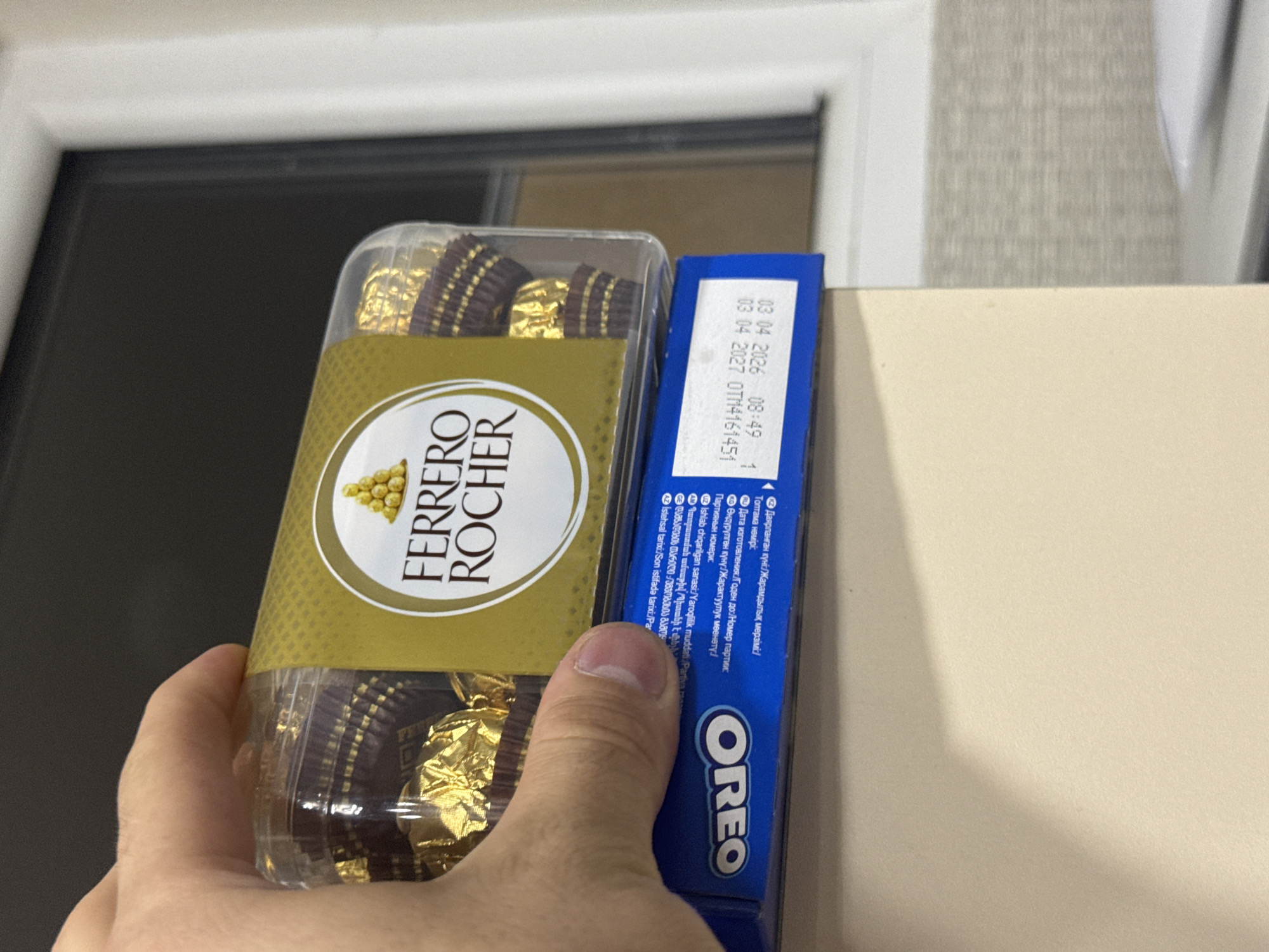 A hand is holding a clear plastic container of Ferrero Rocher chocolates. The container is placed on top of a blue Oreo cookie box. The Oreo box has text printed on it, including an expiration date that reads "03 04 2026". The scene appears to be indoors, possibly on a desk or table, with a blurred background that suggests a window or wall. There is no indication that this image was taken in Tbilisi, Georgia.