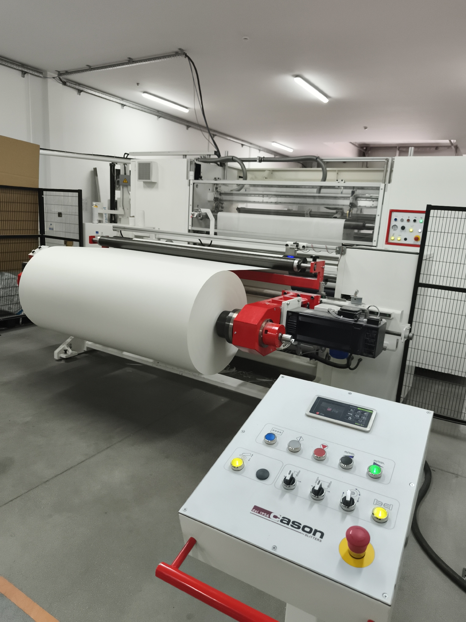 In an indoor industrial setting, a large roll of white material, possibly paper or fabric, is mounted on a sophisticated slitting machine. The machine features prominent red and black components, including motors and rollers, designed for precision cutting. A control panel, positioned in the foreground, displays various buttons, knobs, and a small digital screen with an interface labeled "Cason - SLITTERS". The environment is brightly lit by overhead fluorescent lights, and the floor is a smooth concrete surface. A wire cage fence is visible in the background, suggesting a manufacturing or processing facility. There are no people present in the image.