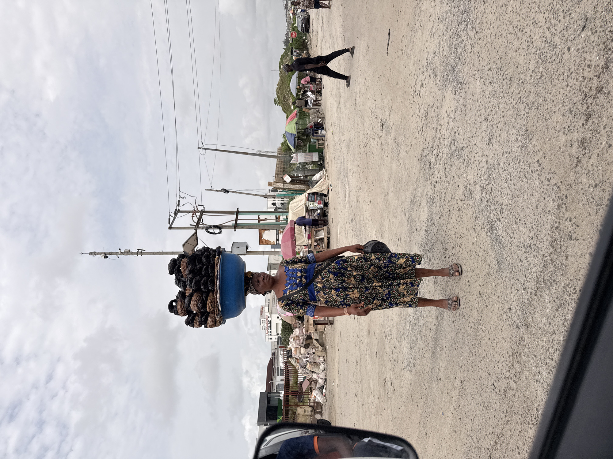 A medium-shot, eye-level outdoor photograph depicts a woman carrying a blue basin filled with what appears to be coiled, dark-colored items on her head while walking on a concrete road. The location is Lekki, Nigeria.
The woman is wearing a patterned blue and gold dress with sandals. She is walking towards the right side of the frame. The basin on her head is filled with many bundles of what appear to be coiled ropes or similar objects.
In the background, there is a bustling street scene with other people, structures with corrugated metal roofing, and overhead electrical wires. A person is walking in the opposite direction, further down the road. A partial view of a vehicle's side mirror and dashboard is visible in the bottom right corner of the frame.
The sky is overcast with gray clouds, suggesting it's either early morning, late evening, or a cloudy day. The road surface is concrete, showing signs of wear.