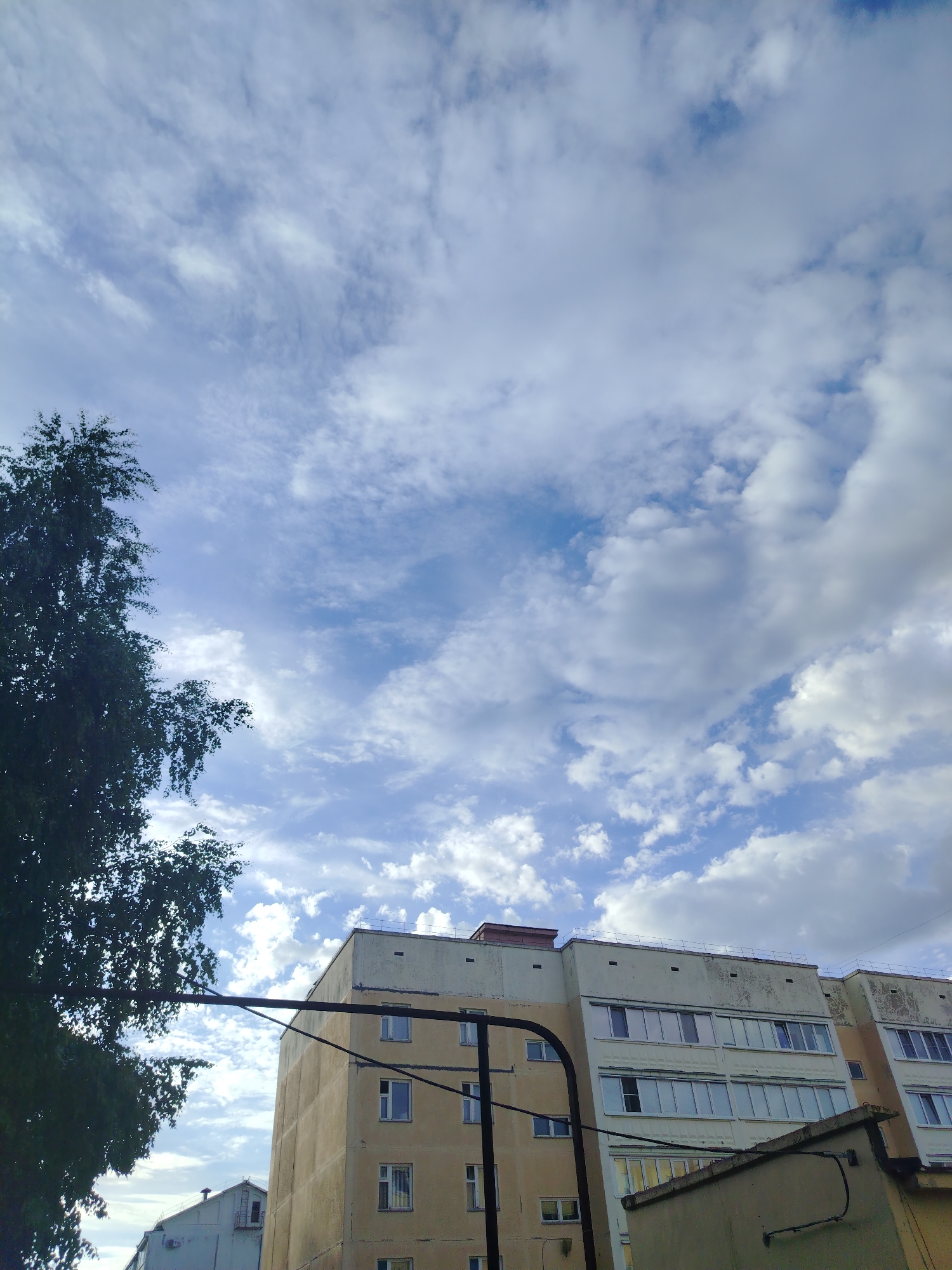 The image shows a multi-story building in Noviy, Russia. The building has a weathered beige exterior and a white top section with windows. A tree branch is visible on the left side of the frame. Electrical poles and wires run in front of the building. The sky is filled with white and gray clouds, creating a partially cloudy sky.
