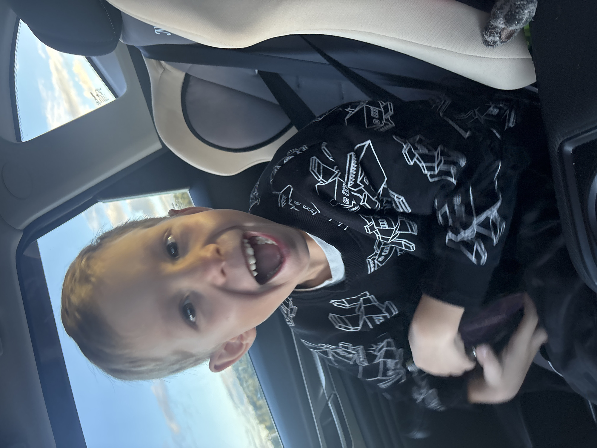 A young boy with blonde hair is sitting in a car seat. He is wearing a black sweatshirt with a white geometric pattern. He is looking at the camera and smiling with his mouth open, showing his teeth. The boy is holding a dark-colored object in his hands. The interior of the car is visible, including a car seat, seatbelt, and part of the dashboard. The windows of the car show a blue sky with some clouds and a hint of green landscape outside. The scene appears to be captured during daylight. No specific location information is provided in the image or crops to determine if it is in Katyshevo, Russia.