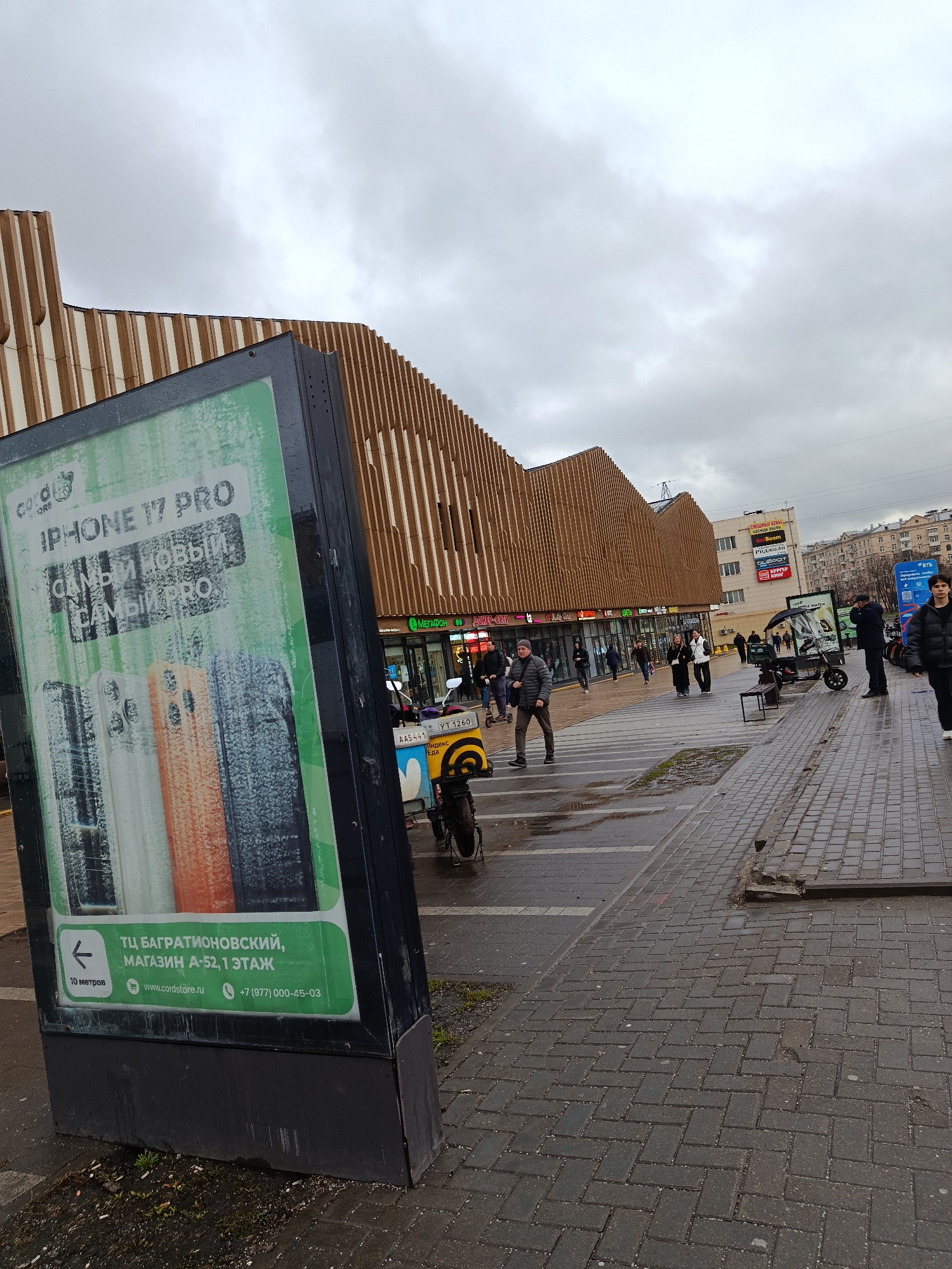 The image shows an urban street scene in Moscow, Russia. A large advertisement for an "iPhone 17 Pro" stands prominently in the foreground, advertising a store called "Cordstore" at the "Bagrationovsky" shopping center. The street is paved with interlocking bricks, and the area appears slightly wet, suggesting recent rain.
A multi-story building runs along the background, featuring a facade with parallel, vertical wooden slats. Commercial storefronts are visible on the ground floor, including a "MegaFon" store and another with Russian lettering. Pedestrians, mainly dressed in dark winter clothing, walk along the sidewalk. A motorized delivery bike is parked nearby. Further back, a building is identifiable with multiple advertisements for retail businesses. The sky is overcast with grey clouds.