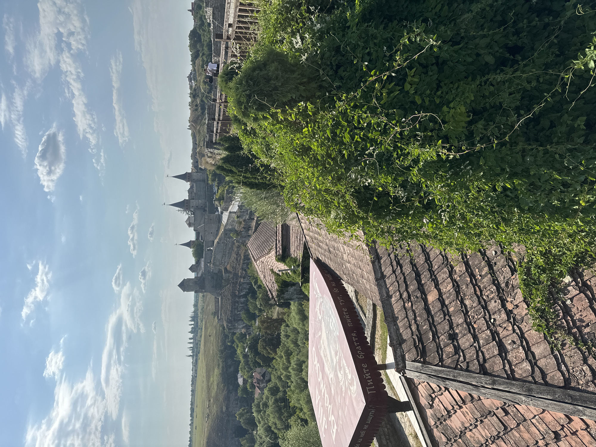 The image captures a panoramic view of the Kamianets-Podilskyi Castle in Kamianets-Podilskyi, Ukraine, on a sunny day with scattered clouds. In the foreground, terracotta-tiled roofs are visible, some showing signs of age and disrepair. A large, red canopy with Cyrillic text is stretched over an outdoor seating area. To the right, dense green foliage covers a significant portion of the frame, with a sculpted evergreen tree standing out.
Beyond the immediate foreground, the historic Kamianets-Podilskyi Castle complex dominates the mid-ground. Its stone walls and multiple towers, including those with conical roofs and flagpoles, are clearly discernible. The castle is situated on a rocky outcrop, overlooking a lush, green valley with scattered buildings and dense trees. A wooden walkway or bridge extends from the castle walls into the distance, with a few individuals walking along it. The background features a gently rolling landscape under a bright blue sky with white, wispy clouds. The overall scene depicts a blend of historical architecture, natural greenery, and a clear sky, suggesting a tourist-friendly location during daylight hours.