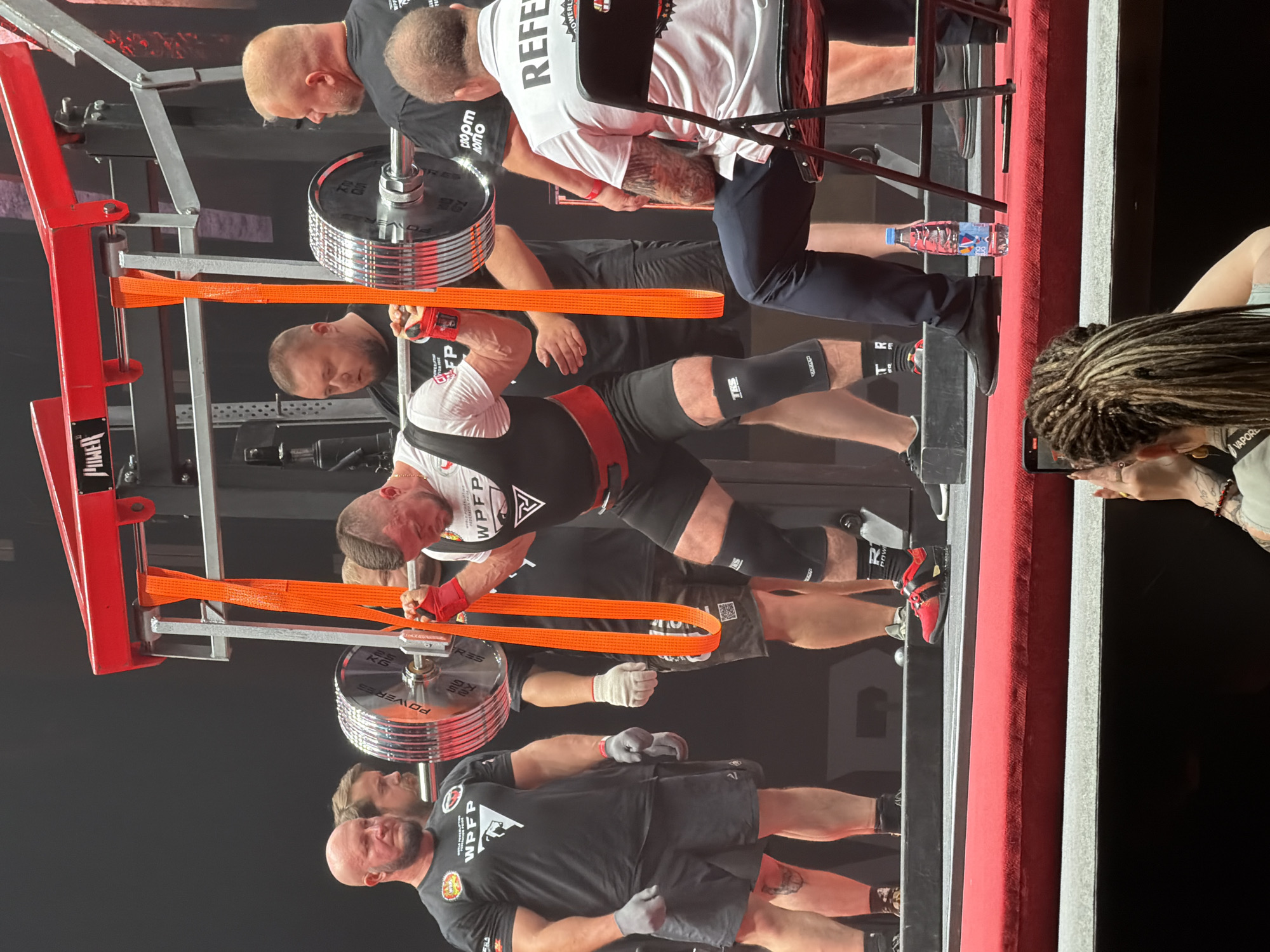 The image depicts a powerlifting competition indoors. A male athlete, wearing a white singlet with black accents, red knee wraps, and black shorts, is in the process of a lift. He is gripping a barbell loaded with large, silver weight plates, attached to an orange strap system. Several other individuals are present around the athlete. To the left, a bald man in a black t-shirt and black shorts stands with his hands at his sides. Behind him, another man in a black t-shirt looks towards the athlete. To the right of the athlete, a man in a black t-shirt with a logo on the sleeve observes the lift. Further to the right, a man wearing a white shirt with "REFEREE" printed on the back is seated on a black folding chair, looking towards the platform. The competition appears to be taking place on a red carpeted stage, with a large metal frame apparatus supporting the barbell. A clear plastic bottle of water is on the floor near the seated referee. The background is dark, suggesting an indoor arena or hall. The overall scene captures a moment of intense physical exertion during a sporting event. The location is identified as the city of Khimki, Russia.