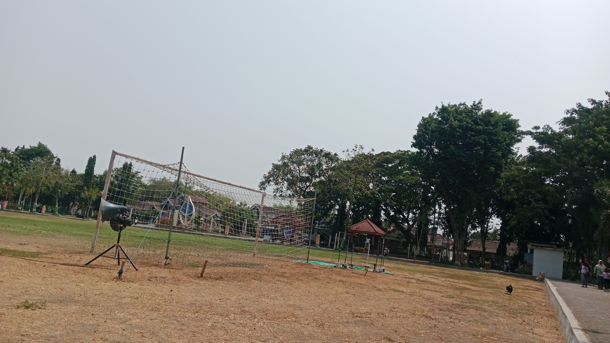 The image depicts an outdoor scene with a sports field in the foreground, featuring a volleyball net and a speaker on a tripod. The field is covered in dry grass and dirt. In the background, there are trees, buildings, and a paved pathway. Several people are visible walking on the pathway. The sky is clear and bright. The scene appears to be a public park or school grounds within the city of Madiun, Indonesia, likely during daylight hours.