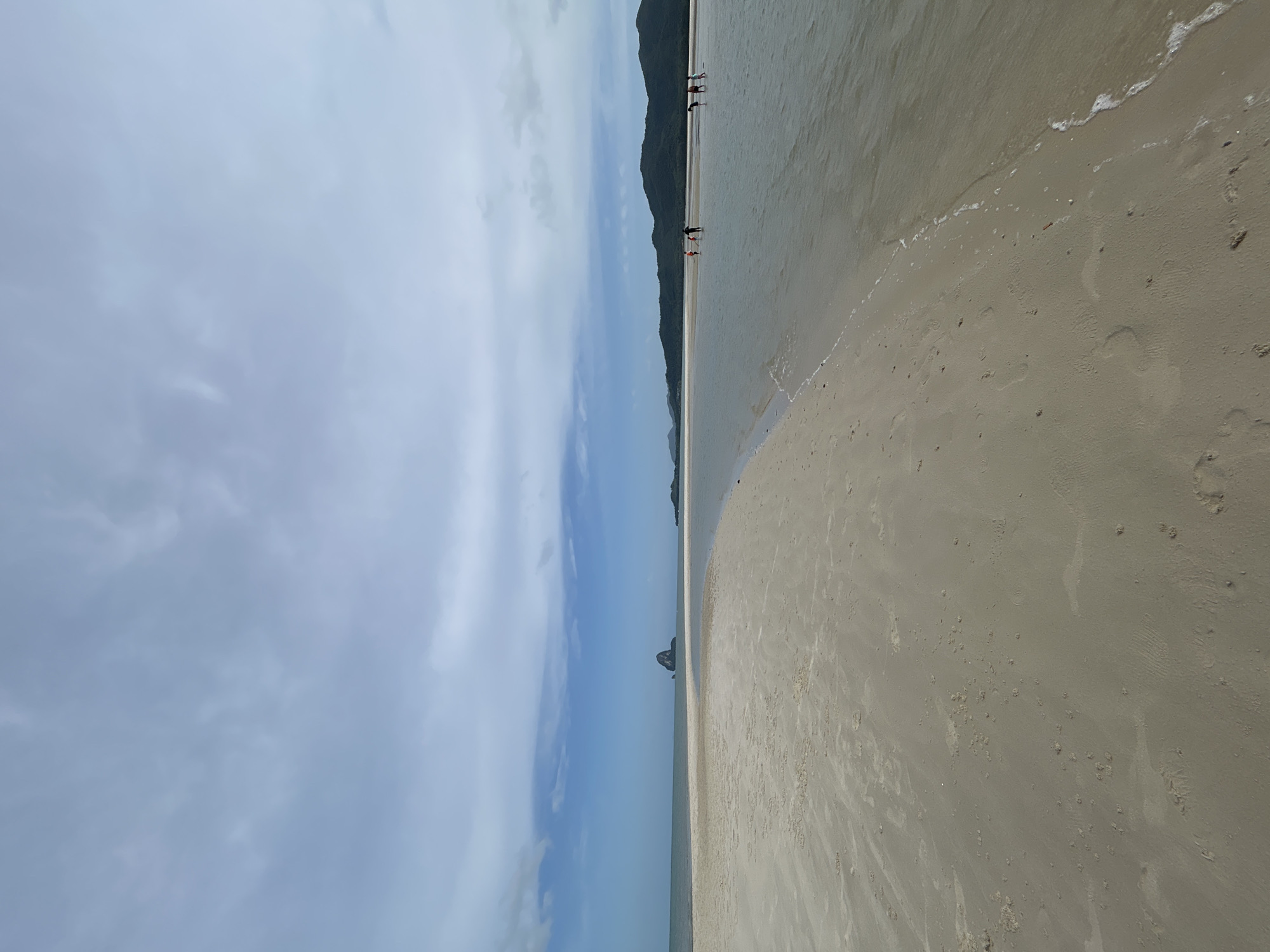 A wide, sandy beach with a shallow body of water stretches across the foreground of the image. The sand is light beige and appears wet in places, with visible footprints and textured patterns. The water is calm and reflects the sky, with gentle waves lapping at the shore. In the distance, a chain of lush, green hills forms the horizon. A distinctive, rocky island with a pointed peak rises from the sea to the left of the frame. Several people are visible in the shallow water and on the distant beach, appearing to be walking or wading. The sky is overcast with a mix of blue and grey clouds. The scene is consistent with the natural beauty of Ko Yao Noi or Ko Yao Yai in Thailand, known for their pristine beaches and unique limestone formations.
