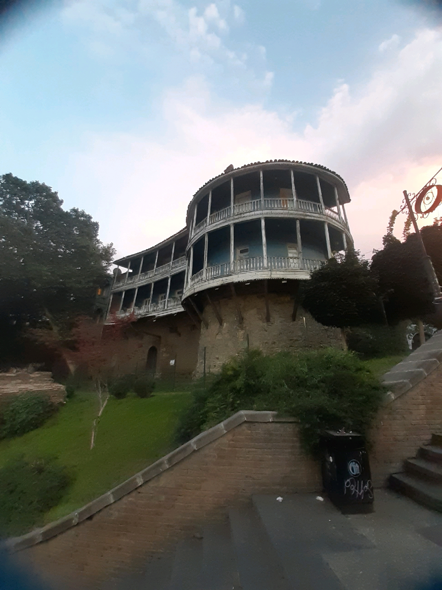 This is a low-angle, wide shot of the historic Abanotubani district of Tbilisi, Georgia, captured at dusk. The scene is dominated by a distinctive, multi-story, circular building with ornate wooden balconies on each level. The building's lower section is constructed of rough stone. Lush green vegetation covers the sloping hillside in front of the building, and a brick pathway leads up to it, bordered by stone steps and a black trash can with graffiti. The sky above transitions from a pale blue to hues of pink and orange, indicating the setting sun. A decorative wrought-iron lamp post with a circular element is visible on the right side of the frame.