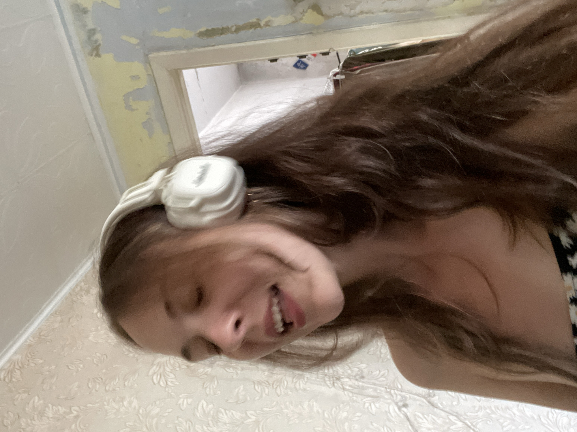 A young woman with long, dark brown hair and closed eyes is shown from the chest up, wearing white over-ear headphones. She is smiling with her mouth slightly open, and appears to be singing or speaking. Her head is turned to the left. The background consists of a wall with patterned wallpaper and a doorway leading to another room, where clothes are visible on hangers. The environment appears to be indoors.